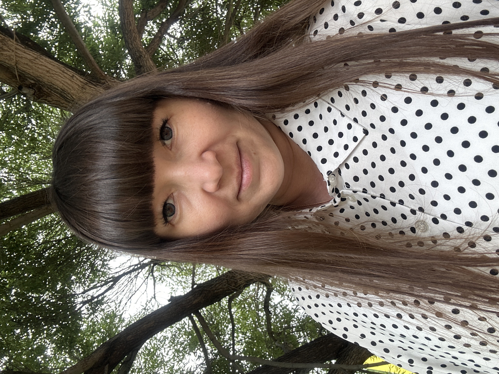 A selfie of a woman is taken outdoors in front of a large tree with visible branches and foliage. The woman has straight, dark brown hair with bangs and is wearing a white collared shirt with black polka dots. She is looking directly at the camera. The background suggests an outdoor, possibly park-like setting, characteristic of locations found within the city of Chaikovsky, Russia.