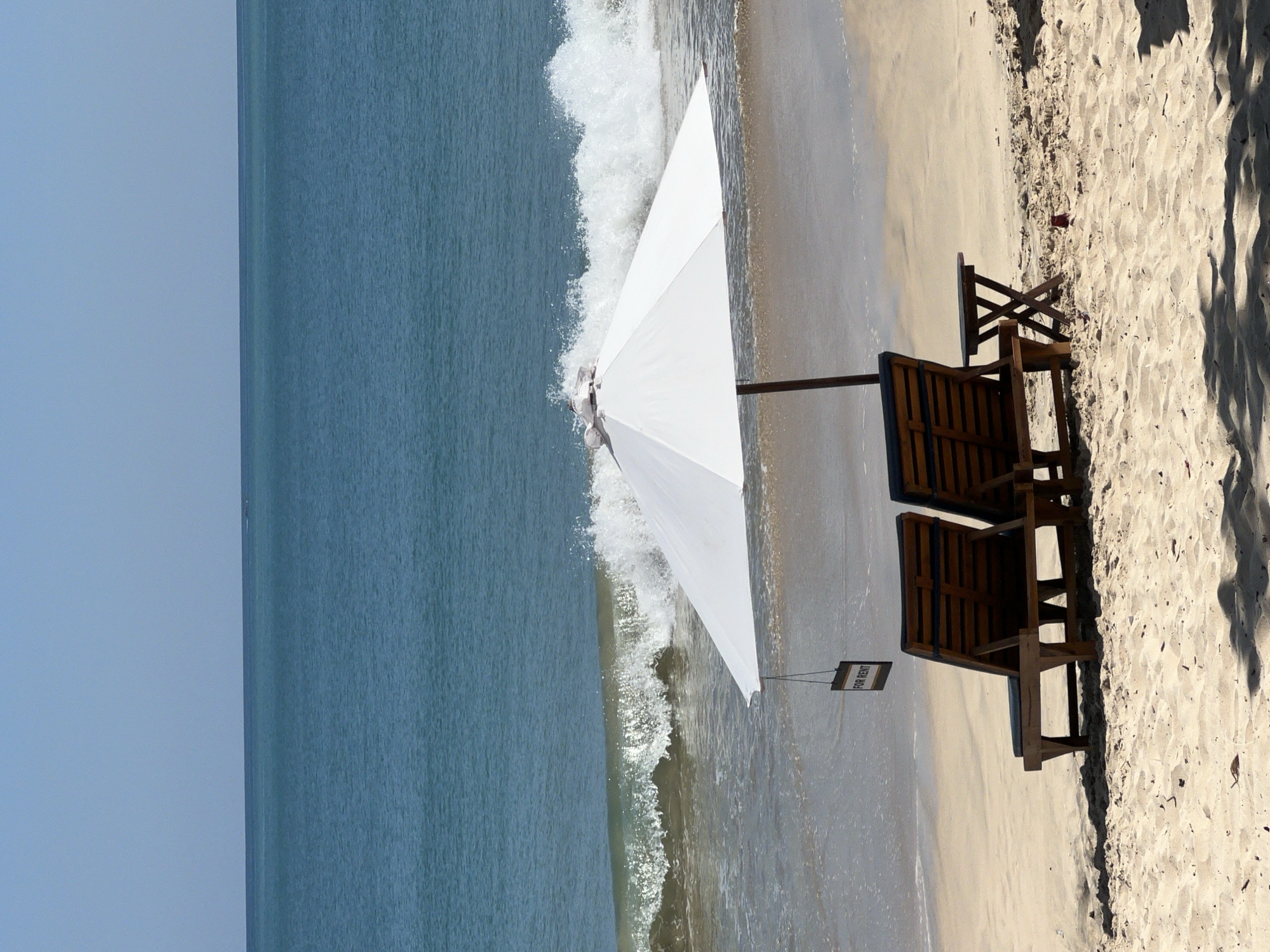The image depicts a beach scene in Kuta Selatan, Indonesia. Two wooden lounge chairs and a small table are arranged under a white umbrella on the sandy beach. A small sign reading "FOR RENT" is attached to the umbrella. The ocean stretches towards the left of the image, with waves breaking near the shore. The sky is clear and blue. Some tree shadows are visible at the edge of the sand.