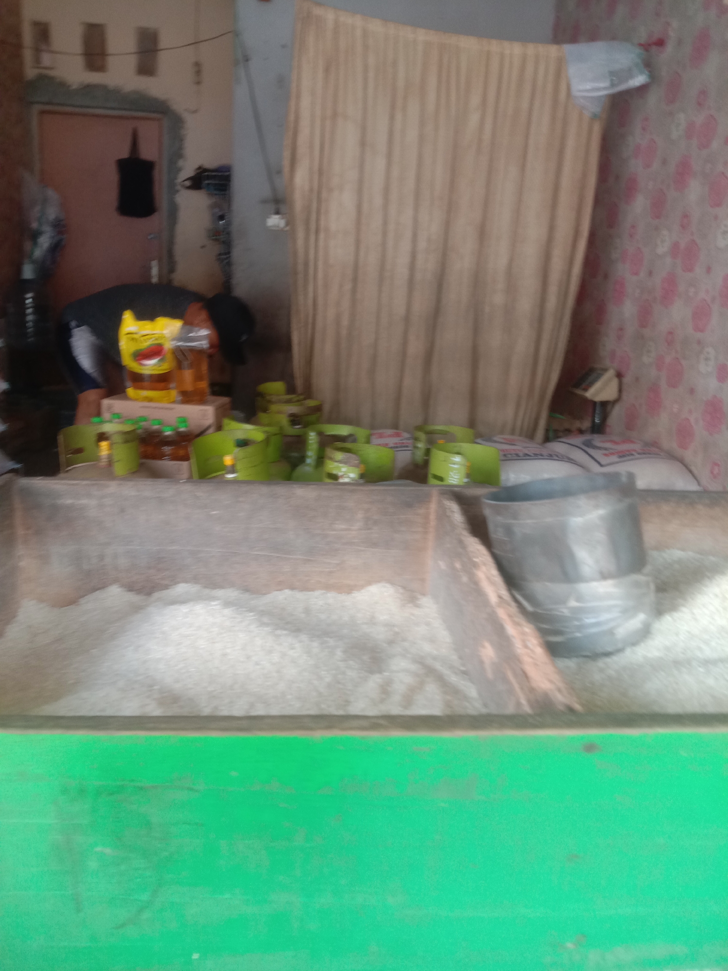 The image depicts an indoor scene, likely a shop or market stall, in Cianjur, Indonesia. In the foreground, a large wooden container is filled with a white granular substance, possibly rice or sugar, which is the primary focus of the lower half of the image. The container has a weathered, rustic appearance and is painted a vibrant green on its exterior base. To the right, a dark gray metal container with a tiered design sits on top of the white substance.

In the mid-ground, a collection of green cylindrical objects, some with visible labels and caps, are arranged on a wooden surface. These appear to be gas canisters, possibly for portable stoves. Alongside them are several clear bottles, some containing amber-colored liquid, likely cooking oil, and some yellow packets with red graphics. A person, partially visible from the back, is seen interacting with these items, wearing a dark shirt and a black head covering. A yellow packet with a cartoon face is prominent among the bottles. Behind these items, large white sacks with red and blue lettering are stacked, suggesting more goods for sale.

The background reveals a wall with a floral pattern, predominantly pink and white. A light brown curtain hangs on the right side of the image, partially obscuring what might be a doorway or window. On the far left, a closed door with a light pink hue is visible, with a black bag hanging on it. The overall lighting suggests an indoor environment with artificial illumination. The scene conveys a sense of commerce, with goods displayed for sale in a somewhat cluttered but organized manner.