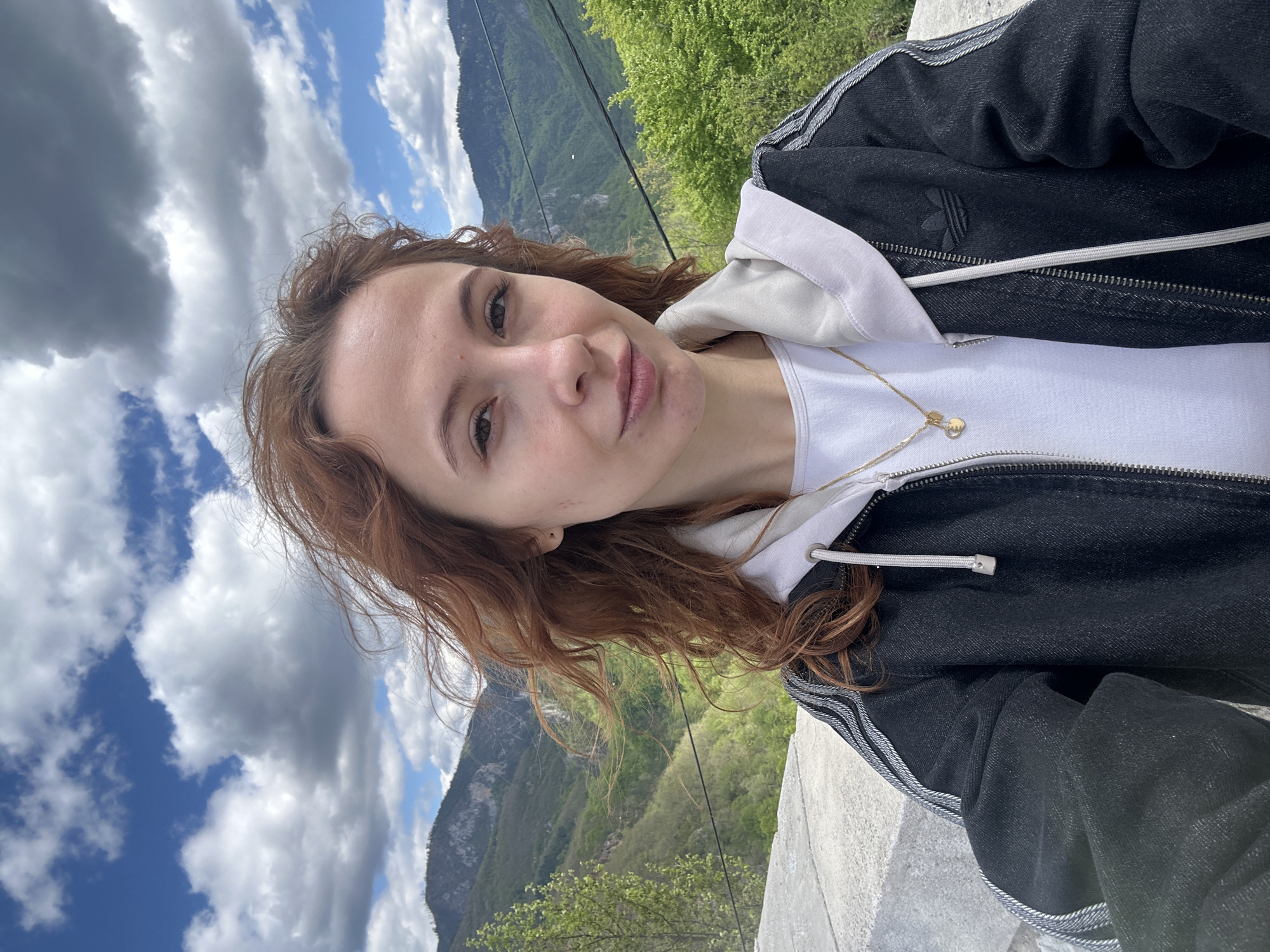 A selfie of a fair-skinned woman with shoulder-length red-brown curly hair. She is wearing a white hoodie under a black Adidas jacket, and a gold necklace. Behind her is a mountainous landscape with trees and vegetation. The sky is partly cloudy with patches of blue. She is in Đurđevića Tara, Montenegro. A gray concrete structure is in the lower right foreground.