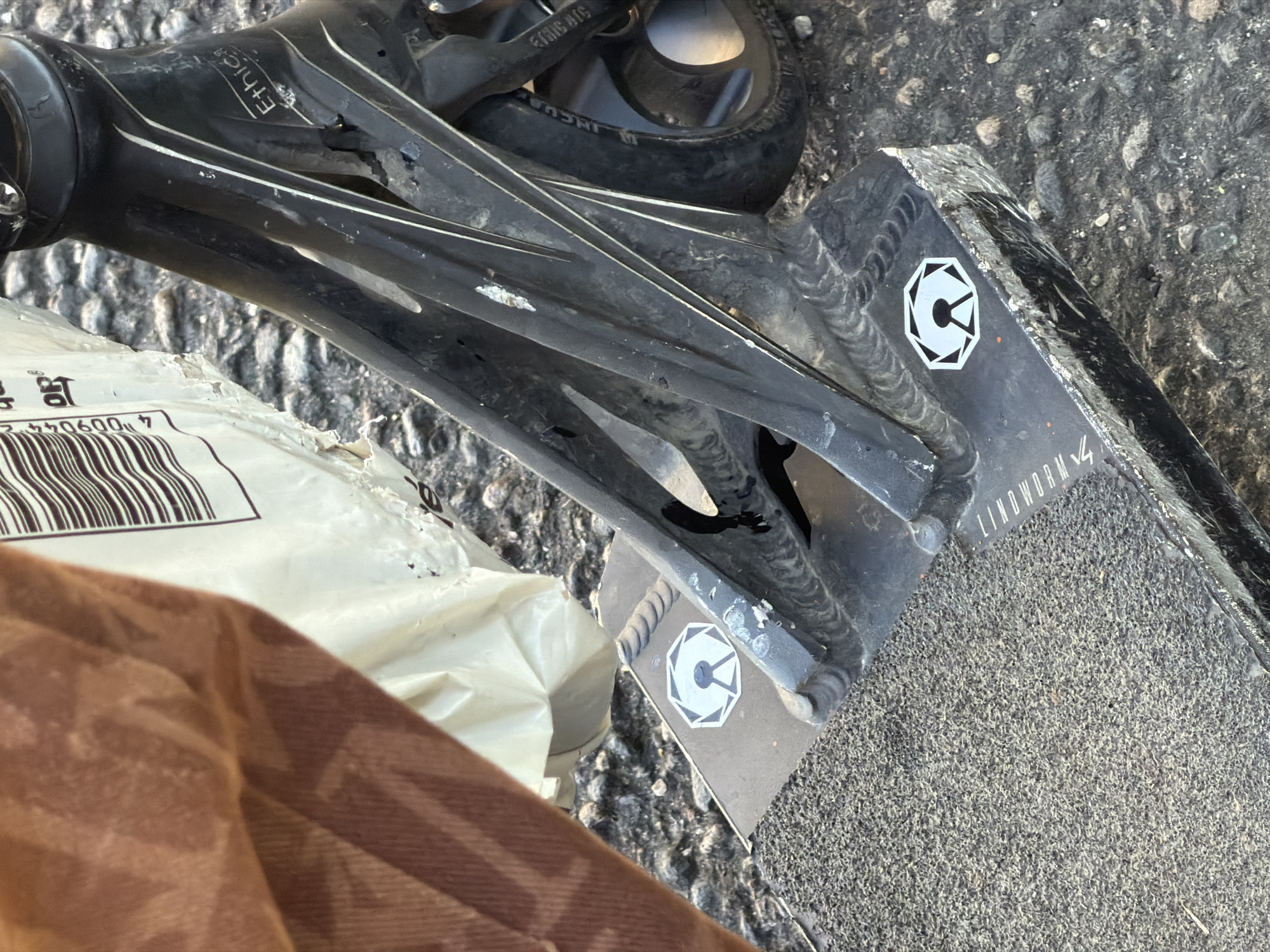 A close-up, low-angle shot shows the rear portion of a black stunt scooter resting on a cobblestone surface. The scooter's deck is covered in grip tape and features two hexagonal stickers with a white geometric design. The metal frame of the scooter is visible, with visible welds and some wear and tear, including scratches and chips. A portion of a brown fabric object, possibly a bag, is draped over the left side of the scooter. In the background, a white plastic bag with a barcode and text is also visible. The lighting suggests an outdoor environment during the daytime. There is no indication of a specific location in Lyon, France.