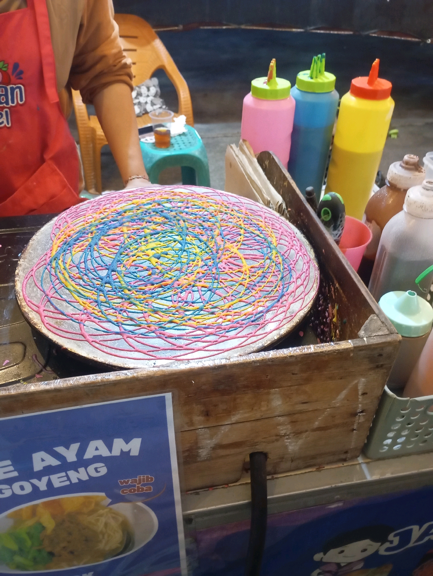 In Babelan, Indonesia, a vendor prepares a colorful dessert on a flat griddle. The dessert, a thin pancake, is decorated with a intricate swirl of pink, blue, and yellow batter. In the background, three squeeze bottles filled with brightly colored batter – pink, blue, and yellow – stand ready for use. To the right, several other bottles, likely containing sauces or toppings, are arranged. A wooden crate holds some of these bottles and appears to be part of the food stall's setup. A red apron, worn by the vendor, is visible on the left side of the image, along with a portion of a light brown shirt. A yellow plastic chair is also in the background, adding to the casual atmosphere of the outdoor stall. A blue sign with white text, partially visible at the bottom, advertises "AYAM GOYENG" (fried chicken).