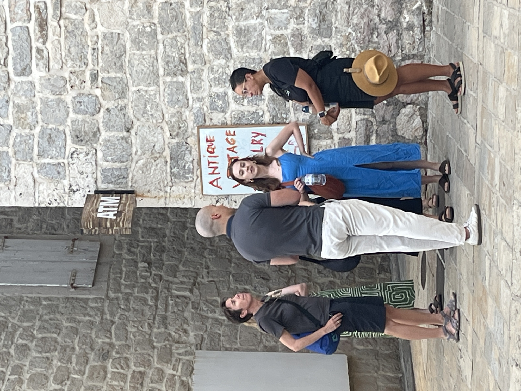 In the city of Budva, Montenegro, four individuals are gathered on a cobblestone street. A sign indicating "ATM" is mounted on a stone wall to the left, above a shuttered window. To the right, a whiteboard with the words "ANTIQUE VINTAGE JEWELRY" is visible.
The person on the far left, a woman with short, dark hair, wears a dark grey t-shirt and dark shorts, carrying a blue bag over her shoulder. Next to her, a man with a shaved head is dressed in a dark grey polo shirt and light-colored pants. A woman in a bright blue dress, holding a water bottle and a brown purse, stands to the right of the man. The fourth individual, a woman with glasses, wears a black t-shirt and black shorts, carrying a water bottle and a hat hanging from her belt. The background consists of a textured stone wall.