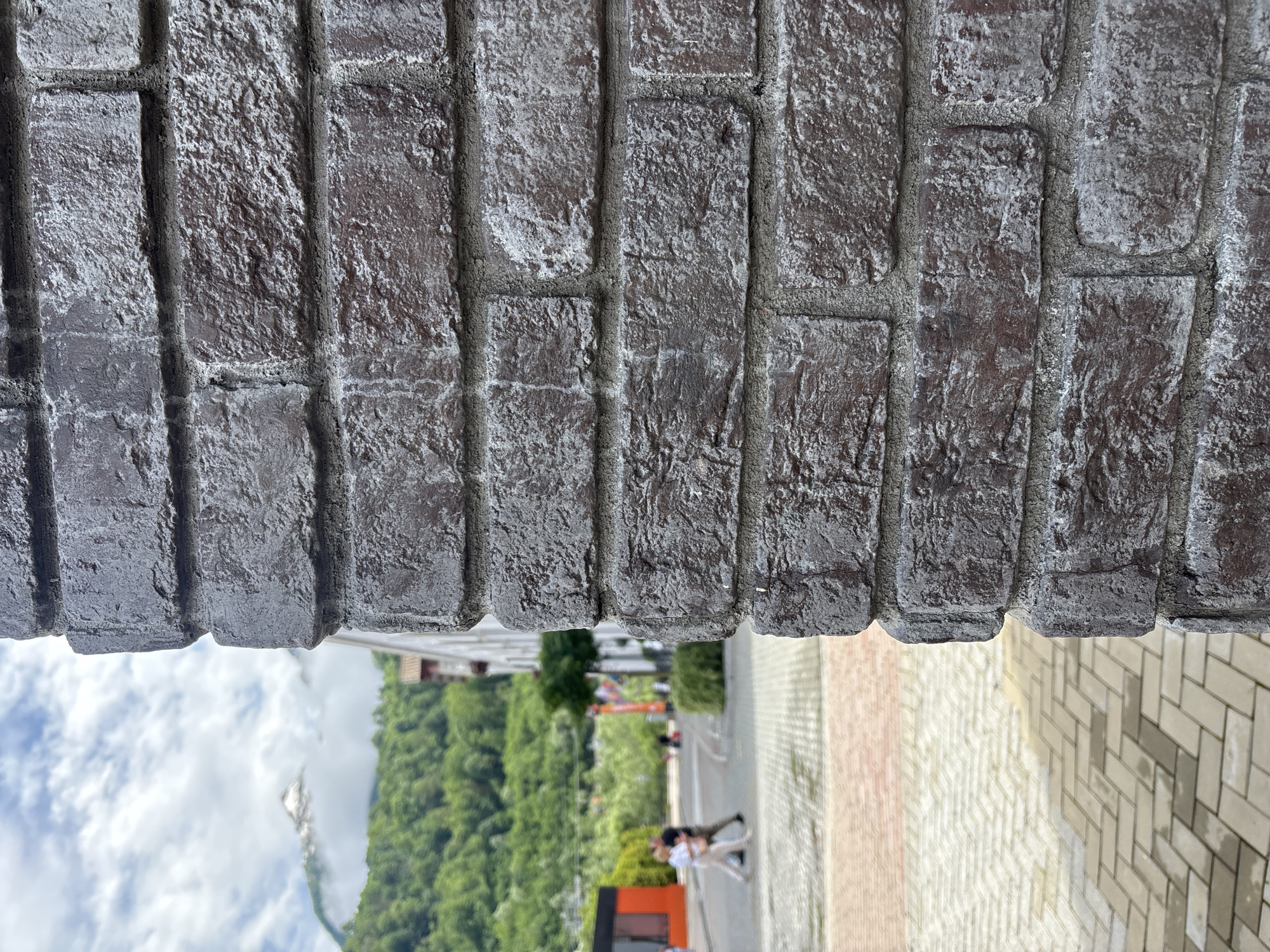 A close-up shot of a dark red brick wall in the foreground dominates the right side of the frame, with its rough texture and gray mortar visible. Beyond the wall, the scene opens up to a street in Estosadok, Russia, under a cloudy sky. A snow-capped mountain peak emerges in the distance, partially obscured by greenery and clouds. Two individuals are walking on the paved sidewalk in the middle ground, one wearing a white top and the other a dark shirt. Adjacent to the sidewalk is a road with a few indistinct figures and structures. Lush green trees and hills form the backdrop, leading to the distant mountain. The paving stones on the ground exhibit a pattern of light and dark rectangular blocks.
