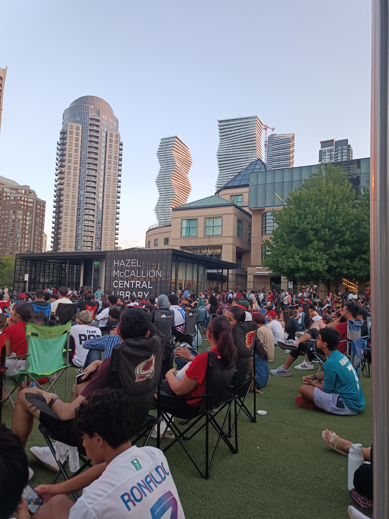 The image captures an outdoor gathering of people sitting on chairs and the ground, facing a building labeled "HAZEL MCCALLION CENTRAL LIBRARY". The scene is set in Mississauga, Canada, as indicated by the building's name, a prominent landmark in the city's downtown core. The background features modern skyscrapers, characteristic of Mississauga's urban landscape. The crowd appears to be watching an event, possibly a public screening or outdoor performance, given their focused attention towards the library building. People are dressed casually, suggesting a relaxed daytime or early evening atmosphere. The overall impression is one of community engagement in a public space, with the library serving as a focal point.