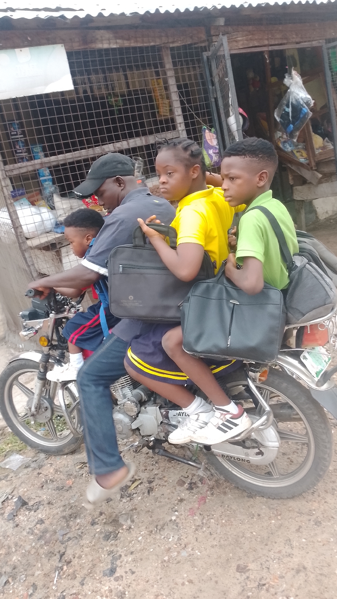 A daytime photograph captures a man transporting three children on a single white and silver motorcycle on a dusty, unpaved road in Lokoja, Nigeria. The man, wearing a black cap, dark blue polo shirt, blue jeans, and light sandals, is operating the motorcycle.
In front of him, a small child in blue and red shorts sits facing forward. Behind the man, a girl with braided hair, wearing a yellow t-shirt and dark blue shorts with yellow stripes, sits holding a black laptop bag. At the very back, a boy in a light green polo shirt and dark shorts sits, also holding a black laptop bag. Another black laptop-style bag is visible between the man and the girl. All three children wear white sneakers, and the yellow stripes on the girl's shorts suggest a school uniform. Their expressions are largely neutral.
The background features simple, rustic shop fronts with corrugated iron roofs and metal grates, suggesting a local market or commercial area. Merchandise, including bottled drinks and other items, is vaguely visible through the grates. The ground beneath the motorcycle is dry and unpaved. The text "DAYLONG" is clearly visible on the rear wheel cover of the motorcycle. The image depicts a common, crowded mode of transportation in the region.