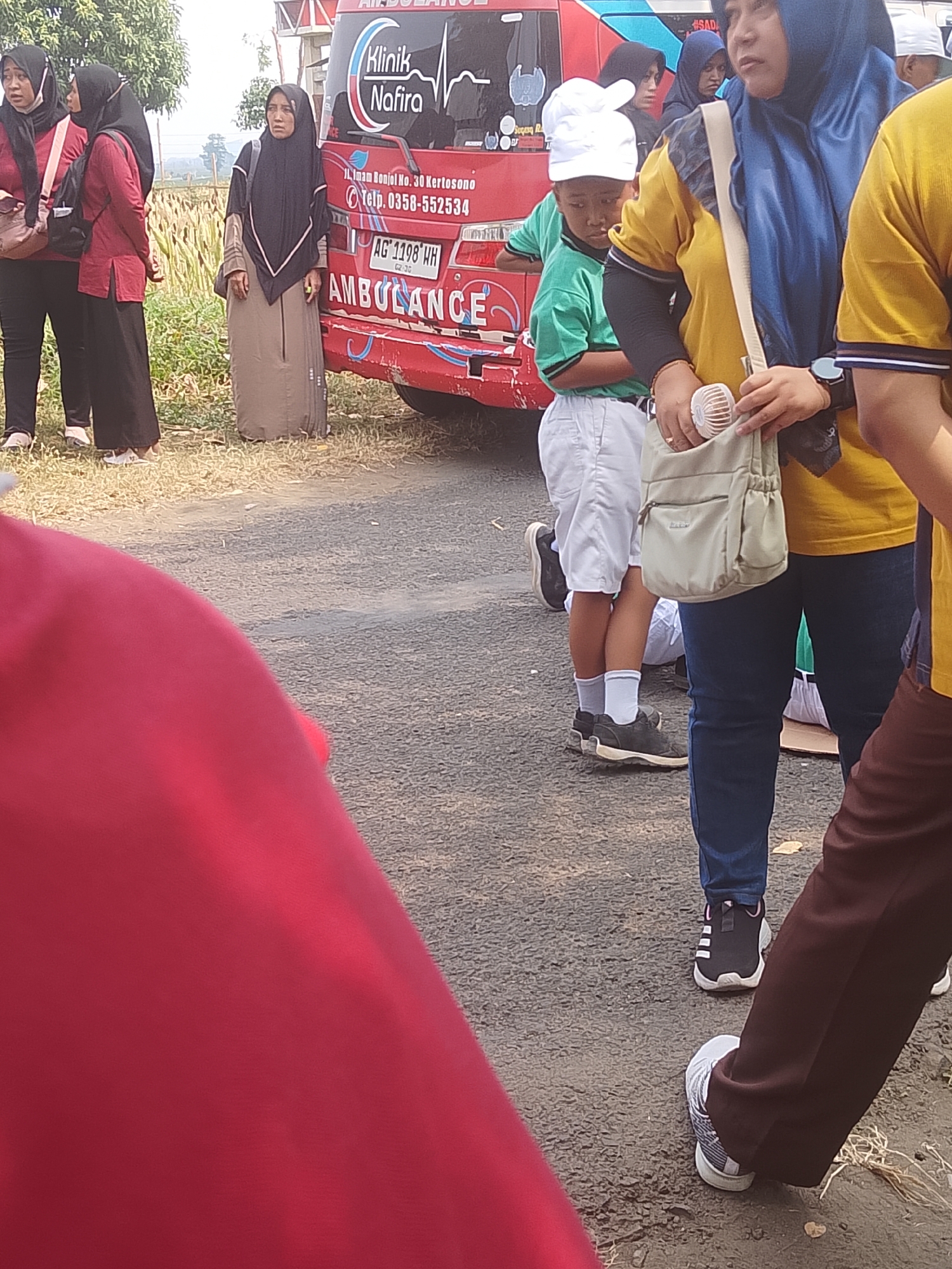 The image shows a group of people standing outdoors near a red ambulance. The ambulance has text on it that reads "Klinik Nafira" and "AMBULANCE". License plate reads "AG 1198 WH". The location appears to be in Bangsri, Indonesia, as indicated by the text on the ambulance. There are several individuals in the image, including women wearing hijabs and children. One woman is holding a small fan in her bag. The ground is paved and appears to be a road or parking area. The background shows some vegetation and trees.