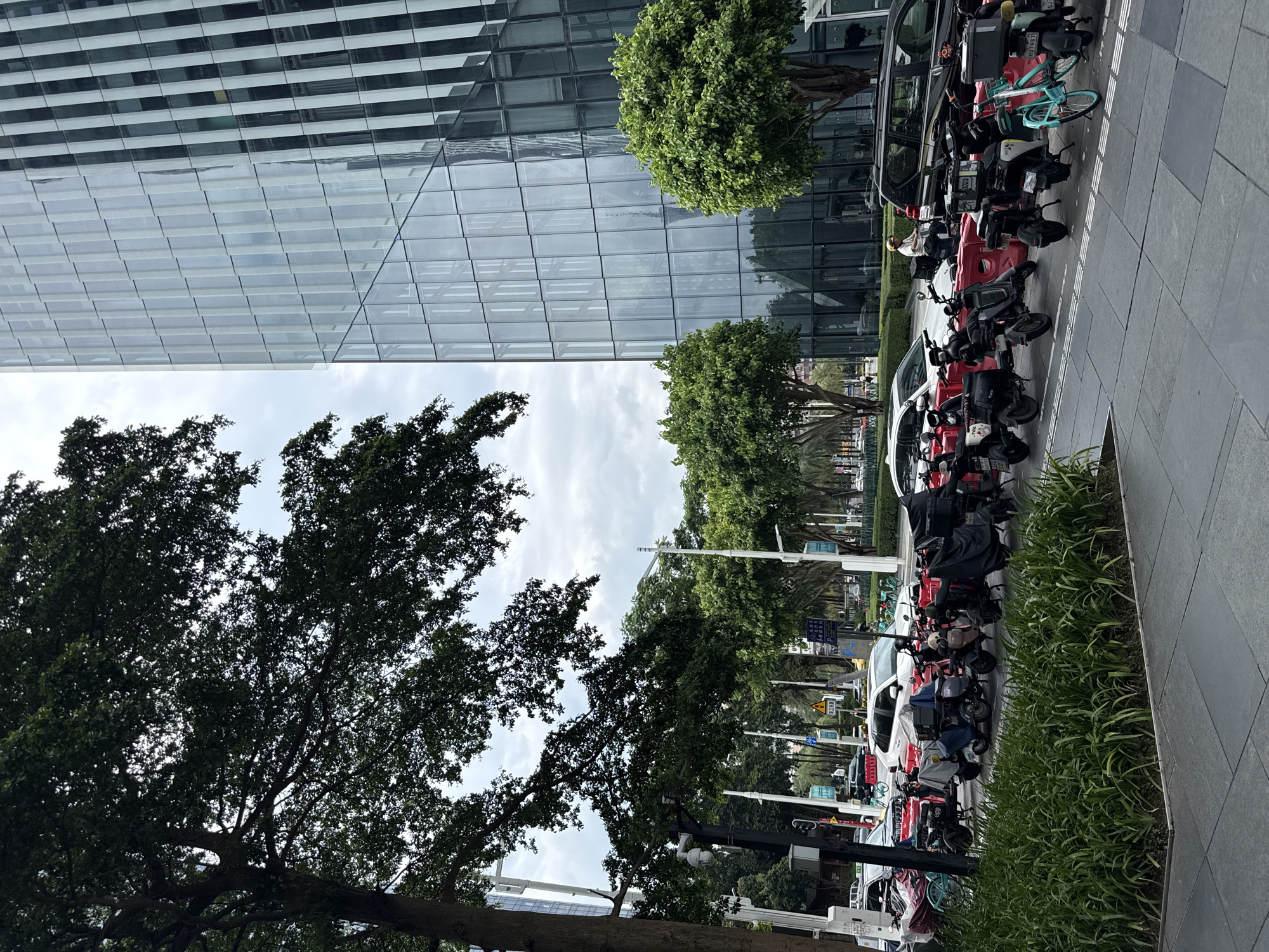 In Tianhe District, China, a street scene unfolds under an overcast sky, dominated by a modern glass skyscraper on the right. In the foreground, a row of parked electric scooters and motorcycles, some covered with tarps, are lined up along a sidewalk. A person wearing a yellow helmet and white shirt is seated on one of the scooters. To the left of the scooters, lush green grass borders the sidewalk. Beyond the scooters, traffic is visible on the street, including cars and a black SUV. Trees line the street, and in the background, a large tree with dark green foliage partially obscures the view of the skyscraper. Streetlights and traffic signals are also present.