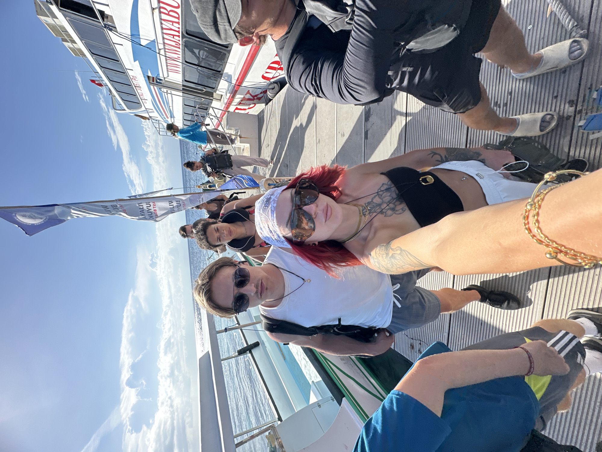 The image shows a group of people on a boat in Nusa Penida, Indonesia. The boat has white and blue paint with red accents and the word "WIJAYABUANA" written on it. Several people are visible in the foreground, including a woman with red hair wearing a bandana and sunglasses, a man with sunglasses, and other individuals. They are standing on a wooden deck with the sea visible in the background. The sky is blue with some clouds.