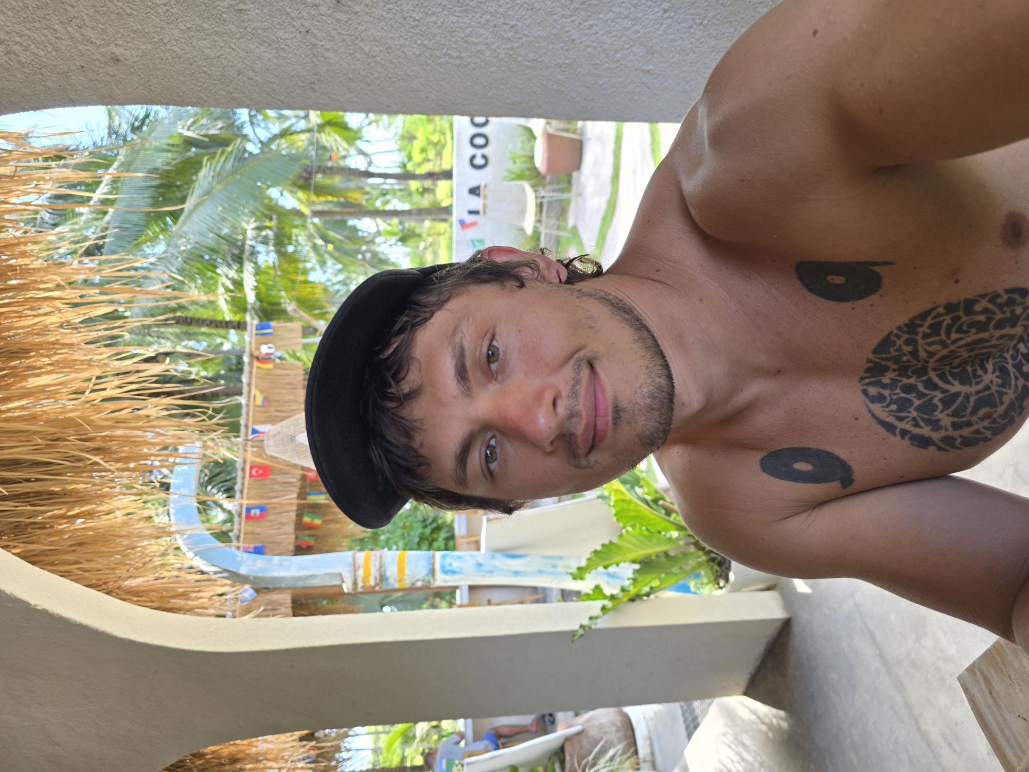 A close-up, tilted, indoor photograph shows a shirtless man with light brown hair and stubble, wearing a black cap. He is smiling at the camera. The man has tattoos on his chest and upper arm. Behind him, in soft focus, are palm trees, flags, and a sign that says "LA COCO". The picture appears to have been taken at La Cocotera Ecolodge in Nusa Penida, Indonesia. Other structural elements are visible, including a roof of thatched material.