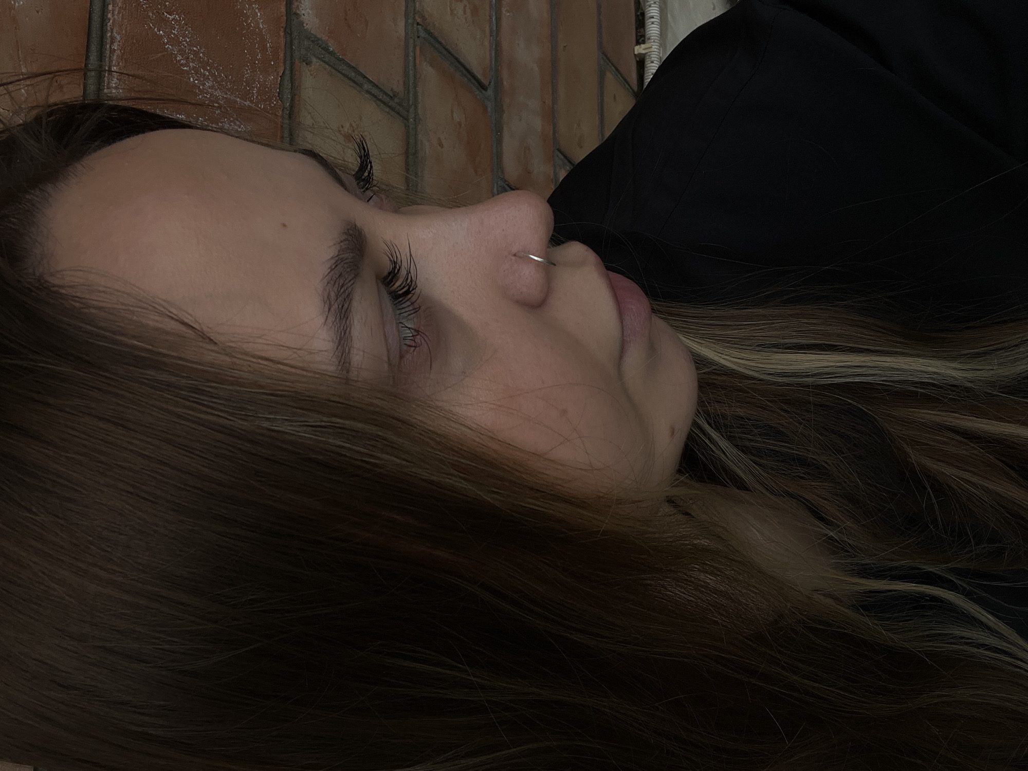 The image shows a woman lying on her side with her head resting near a brick wall. She has fair skin, long brown hair with blonde highlights, and long eyelashes. She is wearing a silver nose ring. She appears to be wearing a black shirt. The location is Novopokrovskaya, Russia. The scene is indoors.