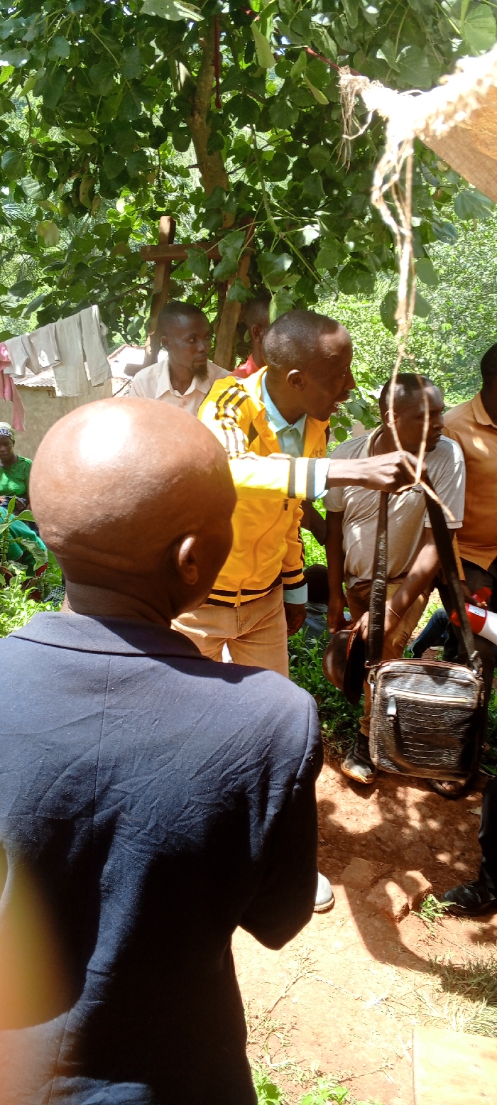 The image depicts an outdoor gathering of several men under the shade of a large, leafy tree in Kundu 1, Democratic Republic of the Congo. The scene is illuminated by bright daylight, casting strong shadows on the reddish-brown dirt ground, suggesting it is midday.
In the immediate foreground, a man with a bald head is seen from behind, wearing a dark blue suit jacket. His back is to the camera. Facing away from the foreground man, a central figure in a distinctive yellow and black striped athletic jacket is actively engaged, holding onto a thick piece of rope or twine that descends from the tree above. He appears to be speaking or demonstrating something to the group.
To the right of this man, another individual holds a dark, textured shoulder bag and a dark hat. Further in the background, other men are visible, observing the interaction. One man behind the central figure has a beard and is wearing a light-colored collared shirt. In the far left background, partially obscured by lush green foliage, a woman in a green outfit can be faintly seen, and clothes are hanging on a line near a simple building structure.
The setting is rural, characterized by dense green foliage, bare earth, and rustic surroundings. The people appear attentive and engaged in what seems to be a community meeting or a discussion. No discernible text is visible on any objects or clothing.