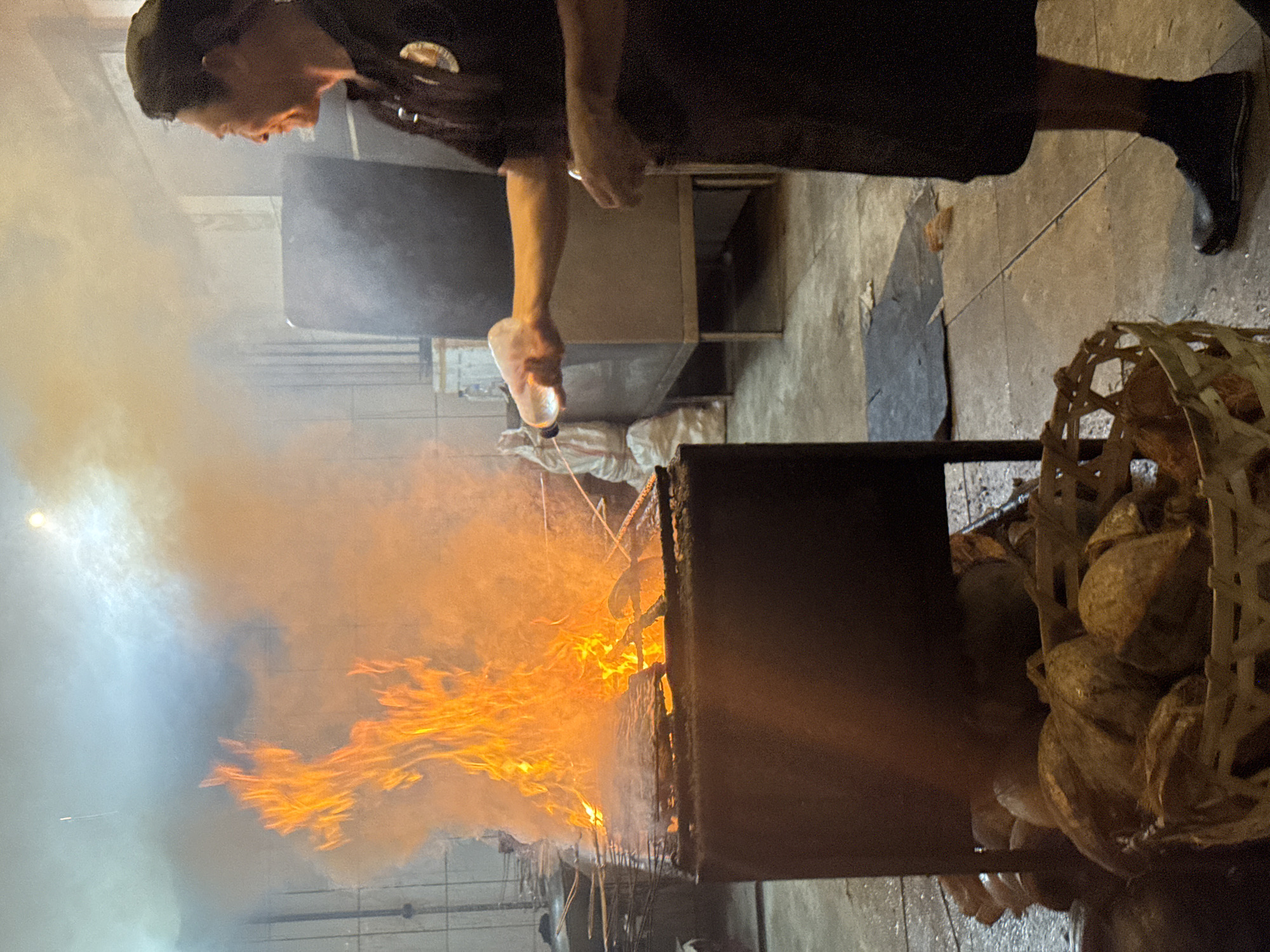 A person in a dark uniform and cap pours liquid from a plastic bottle onto a fire. The fire is contained in a metal structure. Beside the fire is a basket filled with what appear to be coconuts. The location is Kuta Selatan, Indonesia.