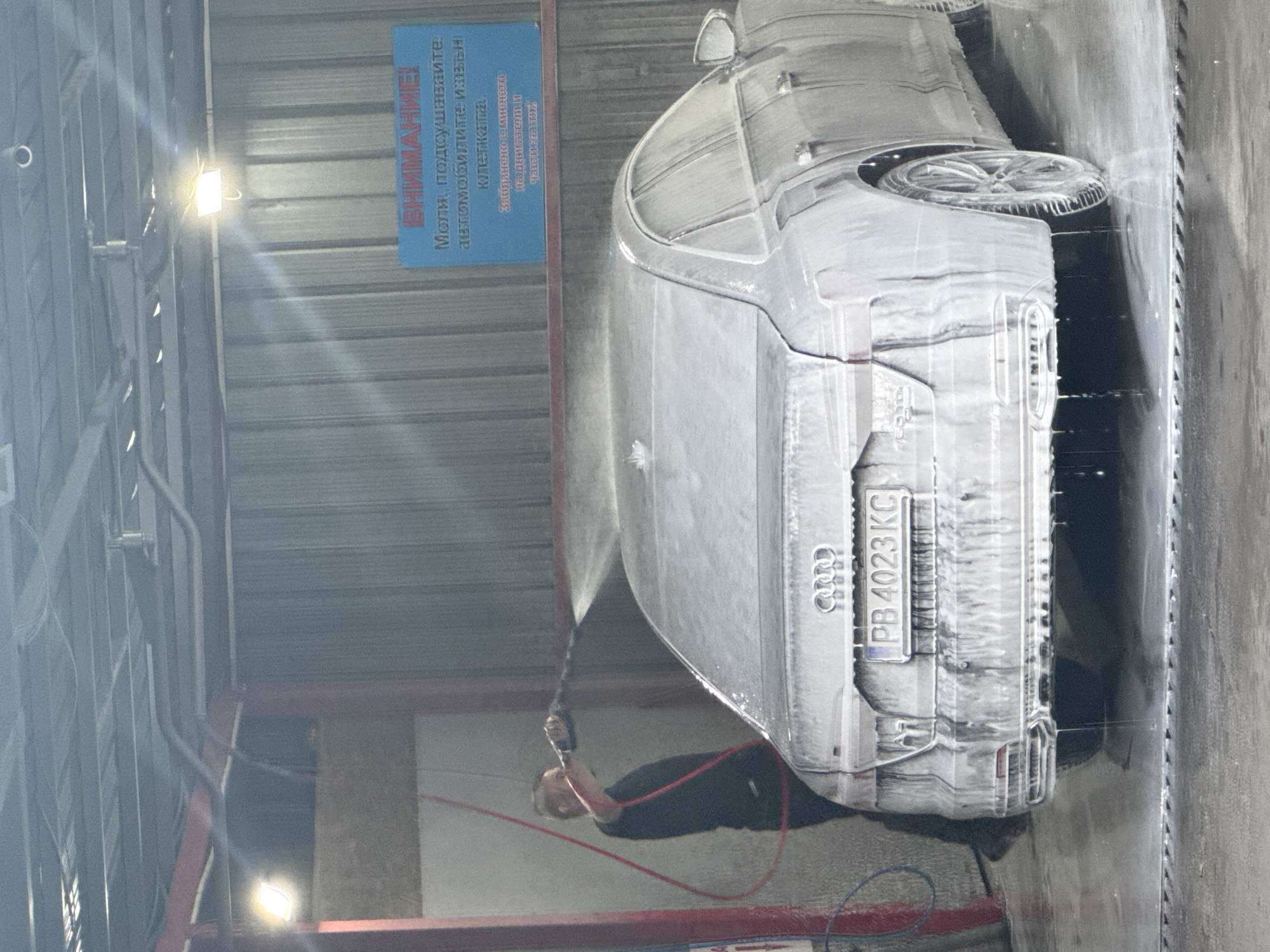 A man is washing a silver Audi with license plate PB 4023 KC at a carwash in Plovdiv, Bulgaria. The car is covered in soap suds. A sign in Cyrillic is visible on the wall, reading "ВНИМАНИЕ! Моля, подсушавайте автомобилите извън котелката." The carwash area appears to be a metal structure with overhead lighting.