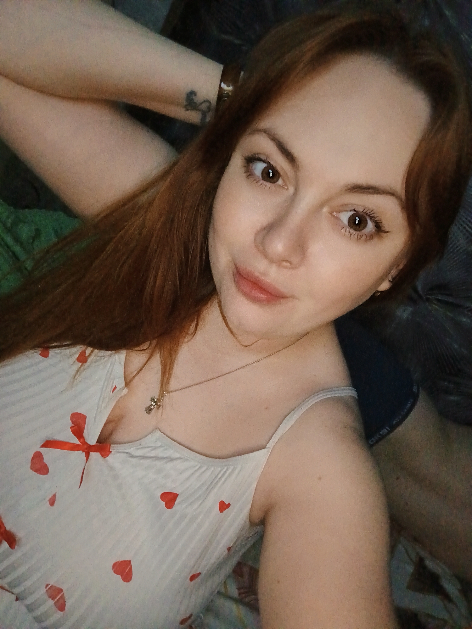 A woman with long red hair is lying down, wearing a white tank top with red hearts and a red ribbon at the neckline. She has brown eyes and light skin. A silver necklace with a cross pendant is visible. Her left arm is raised with a tattoo visible on the wrist. A person is partially visible on the right side of the frame. The location is unknown and there is no evidence it is in Chaikovsky, Russia.