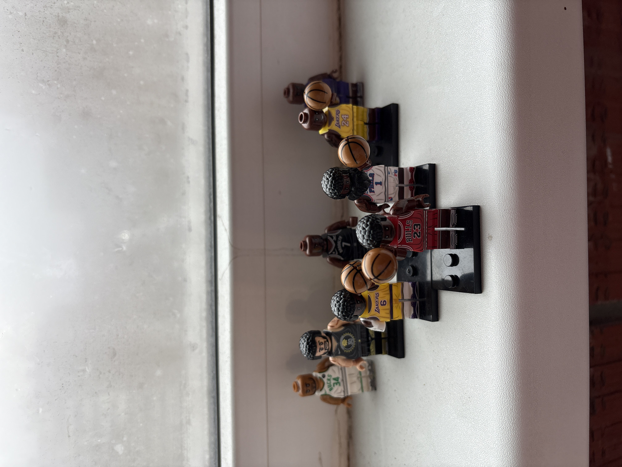 The image shows a row of Lego basketball player minifigures lined up on a white surface, likely a windowsill, next to a window. The minifigures are wearing jerseys with various team colors and numbers, and some are holding basketballs. From left to right, minifigures sport jerseys representing teams like the Celtics (34), Warriors, Lakers (6), Bulls (23), and 76ers (1). The window is slightly frosted or has condensation on it. The location is not discernible as Izhevsk, Russia, from the available visual information.