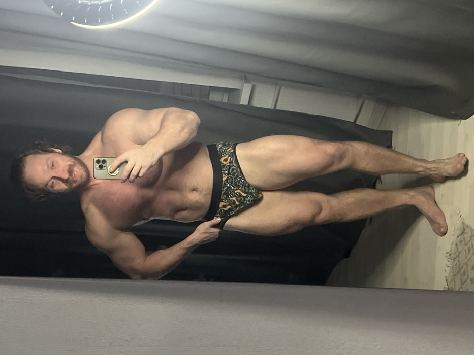 A muscular man poses in front of a mirror, holding a smartphone. He's shirtless, wearing patterned underwear. His physique is well-defined, with visible muscle tone in his arms, chest, and legs. The lighting is somewhat dim, with a light fixture visible in the upper corner. The background features dark curtains or blinds. The man has a beard and appears to be looking at his reflection.