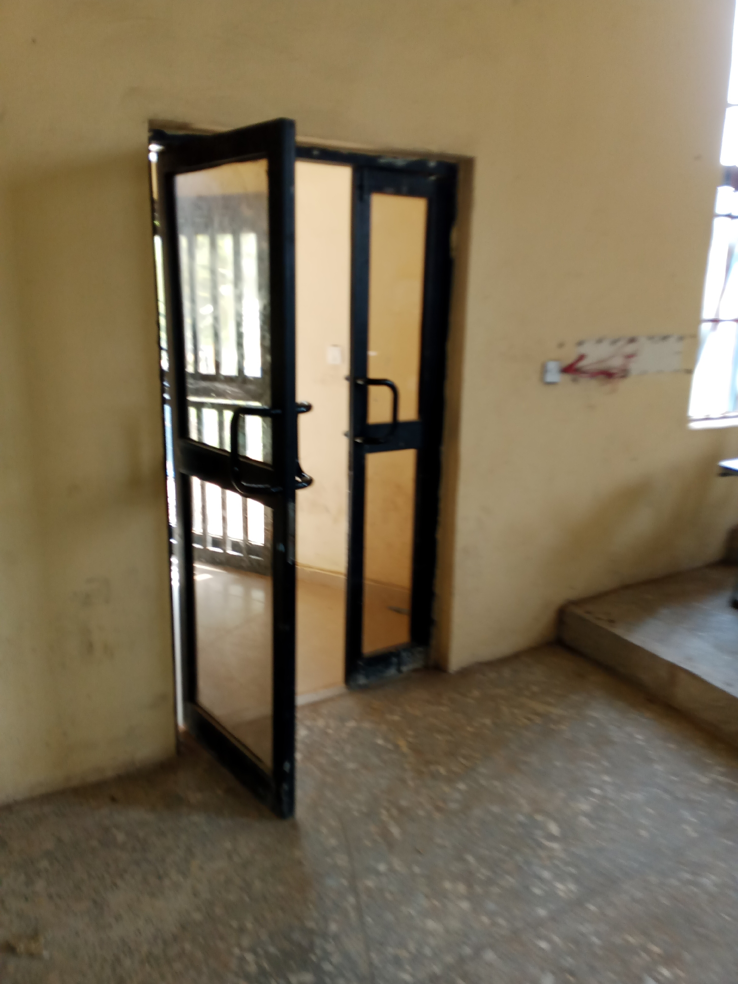 The image depicts an interior scene, likely a hallway or entryway, with a double door slightly ajar. The walls are painted a light, matte yellow, and the floor appears to be concrete with a speckled pattern. To the left, one of the doors, made of black metal and glass, is open, revealing a glimpse of a room with barred windows. The other door, partially closed, also has a black metal frame and glass.
On the wall to the right, a white sign with red markings and an arrow pointing left is visible. Further to the right, a concrete staircase ascends out of view. The lighting suggests an indoor environment, possibly illuminated by natural light from unseen windows or artificial lighting. The overall impression is of a functional, unadorned space. The presence of the staircase and the type of doors suggests this could be within a public building, educational institution, or residential complex. Without further context or identifying markers, it is impossible to definitively pinpoint the specific location within Jalingo, Nigeria.