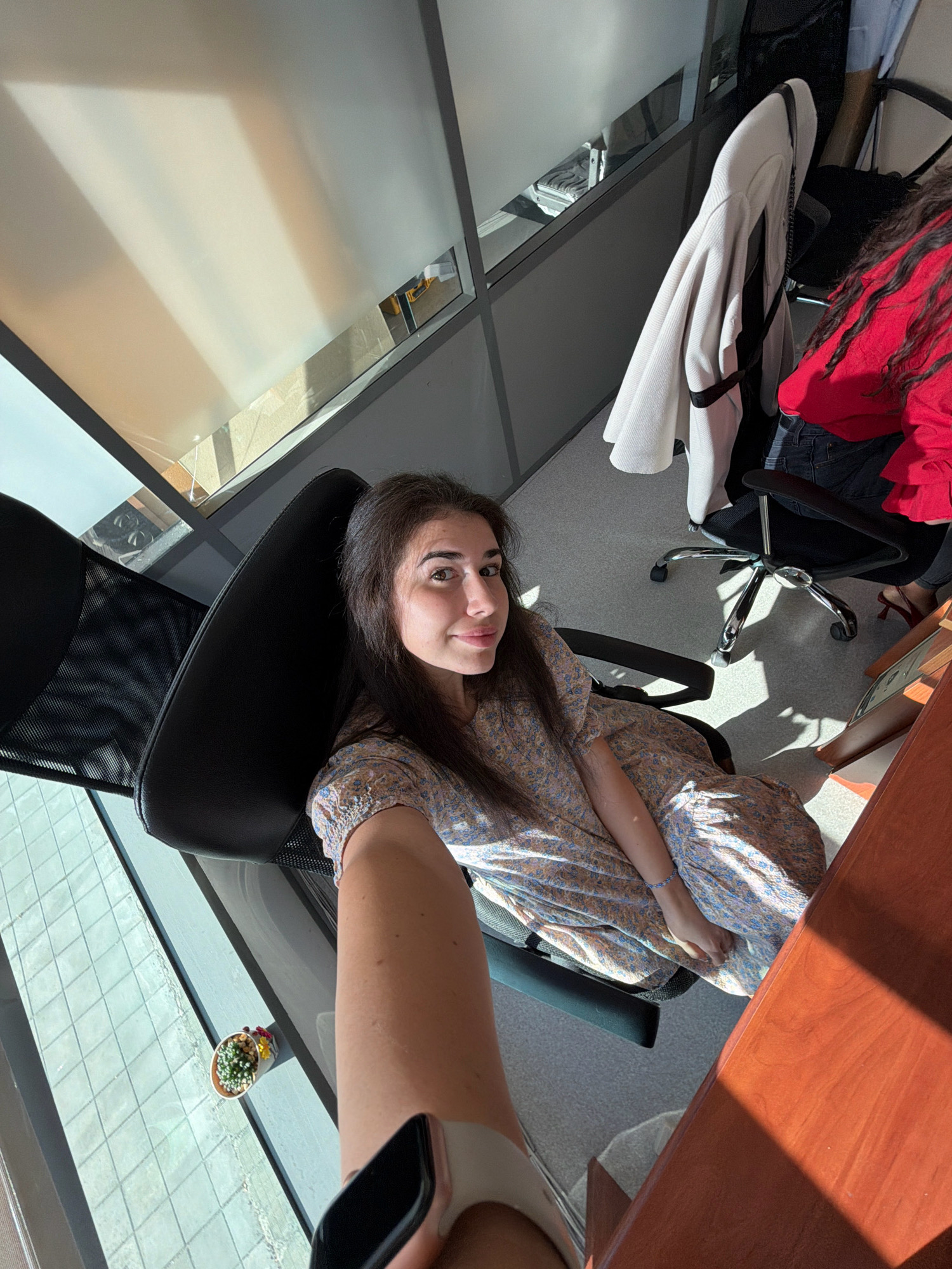 A young woman with long dark hair is seated in a black office chair in a modern office setting. She is wearing a light-colored floral dress and a smartwatch on her left wrist. The office has frosted glass partitions and is illuminated by sunlight streaming through a large window, creating distinct shadow patterns on the floor and wall. In the background, another person in a red top is visible, also seated in an office chair, and a beige jacket is draped over a chair. A small potted plant sits on the windowsill. The location is an office in Sofia, Bulgaria.