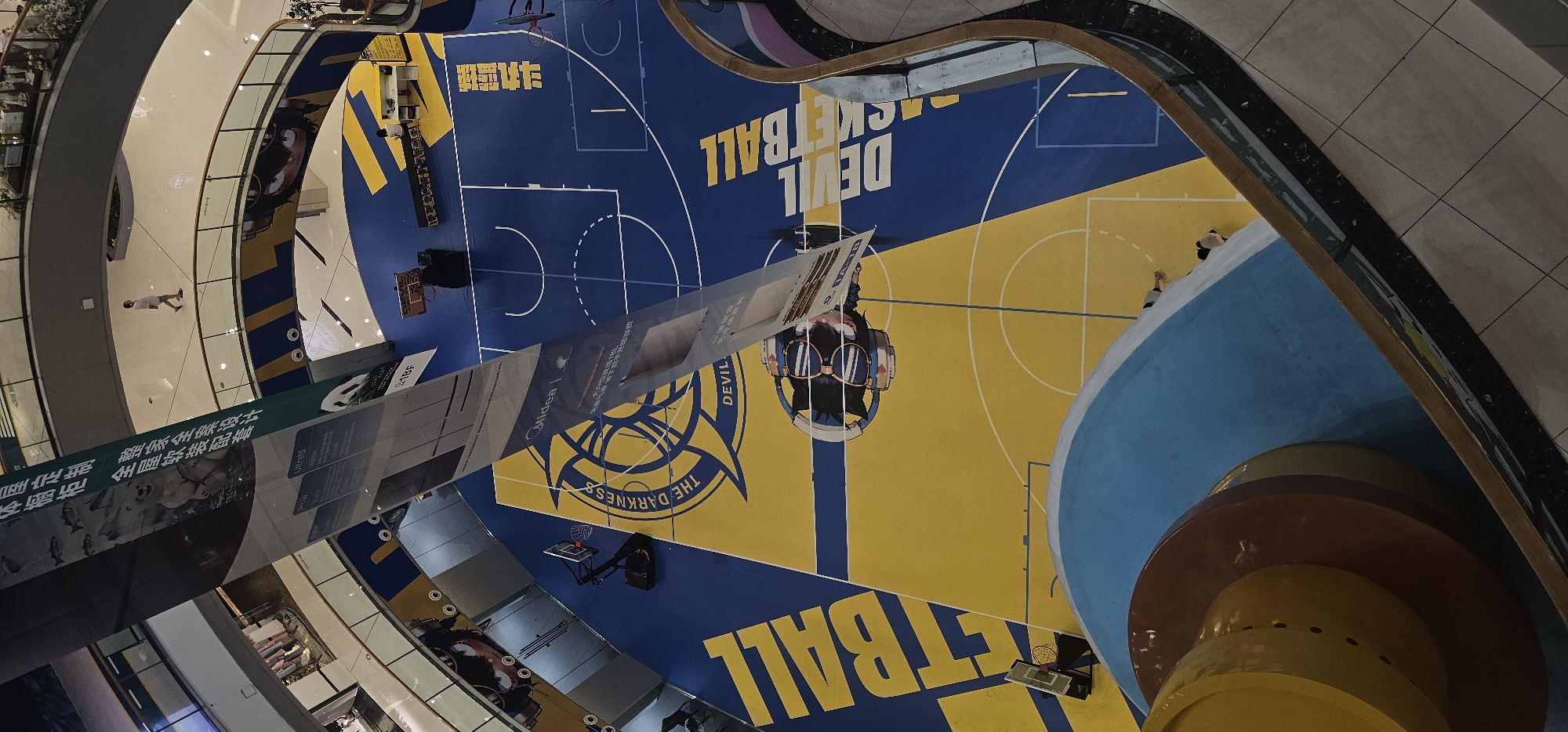 The image captures an aerial view of an indoor basketball court, likely located within a shopping mall in Shapingba District, China. The court features a vibrant blue and yellow design, with markings for a basketball game. A large banner hangs from above, partially obscuring the court and displaying images of animals and text in Chinese. Several individuals are visible on and around the court, including a person walking on a higher level of the mall, a person sitting at a desk on the court, and two individuals appearing to be riding or interacting with a circular, multi-colored structure in the foreground. The environment suggests a commercial or recreational space with multiple levels and walkways.