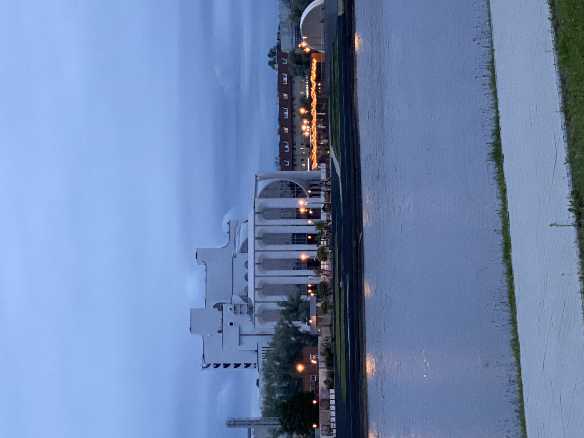 The image shows the Veliky Novgorod Concert Hall, a modern white building with a distinctive curved facade, situated on the bank of the Volkhov River. The sky is overcast and a deep blue, indicating dusk or twilight. Lights are visible in the windows of buildings across the river and along the embankment, reflecting on the water. The foreground shows a paved walkway and some sparse vegetation. The overall atmosphere is calm and serene, with the architecture of the concert hall dominating the scene.