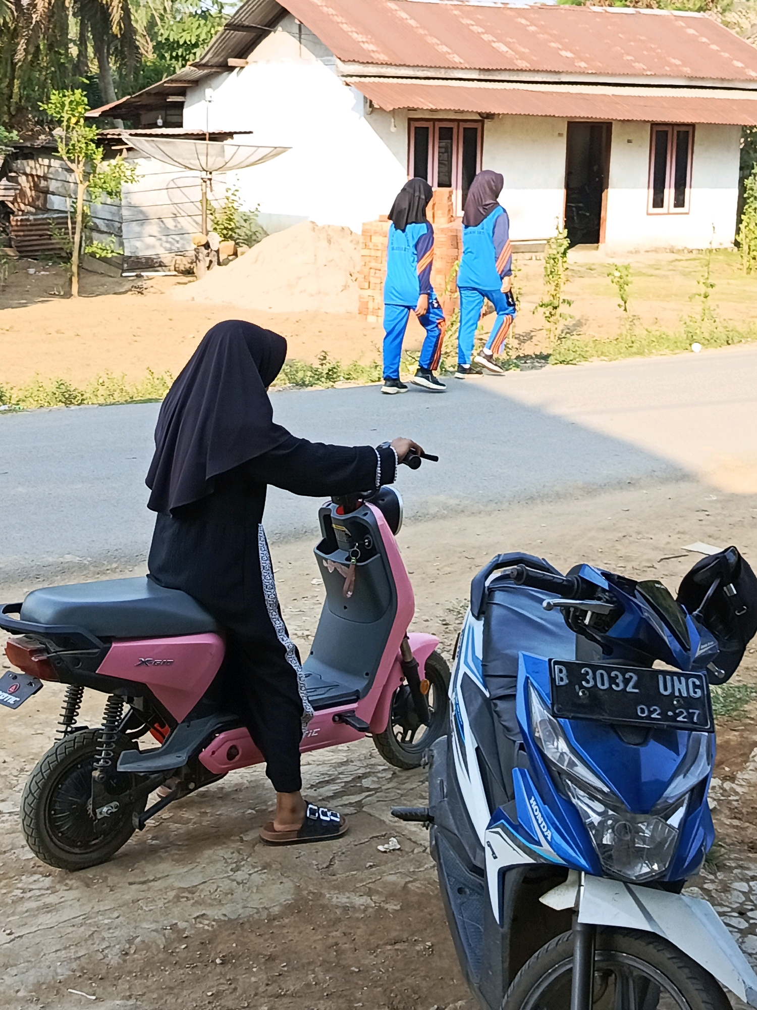 In the city of Megang Sakti I, Indonesia, a person in a black hijab and long black clothing is seated on a pink electric scooter, facing away from the camera. The scooter has the model name "X-Bic" printed on its side. Another motorcycle, blue and white with a black license plate reading "B 3032 UNG 02-27", is parked in front of the pink scooter. In the background, two individuals wearing blue athletic uniforms with orange trim are walking away from a small, white building with a corrugated metal roof. The building has a satellite dish mounted on its side. The environment appears to be a rural or semi-rural setting, with dirt ground and sparse vegetation.