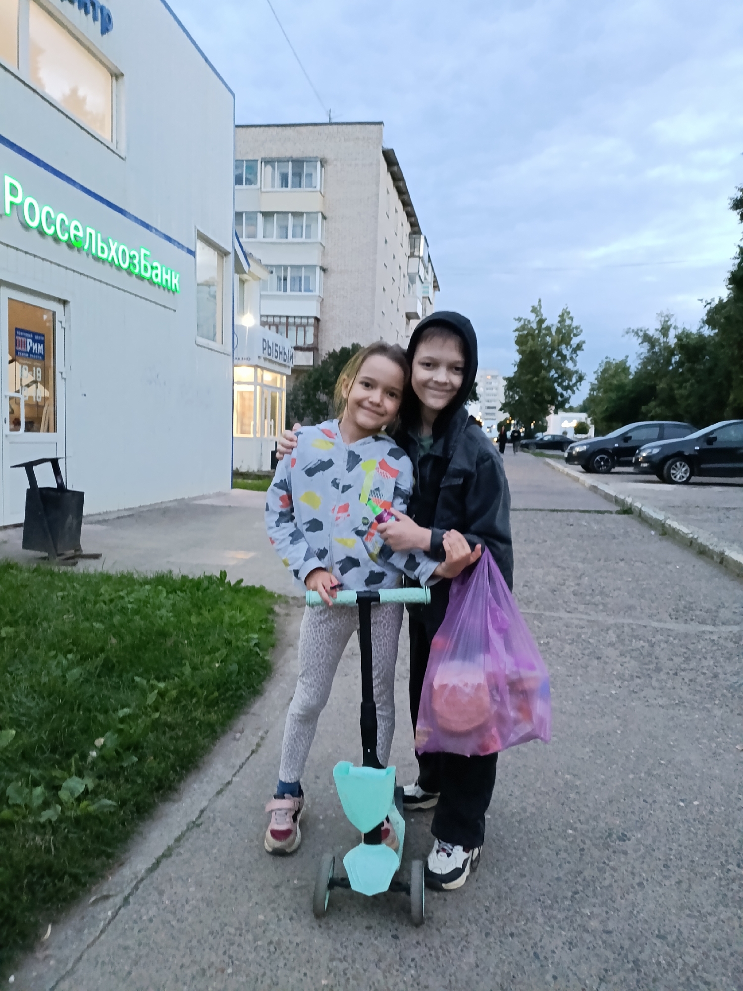 The image shows two young girls standing on a paved sidewalk in front of a building with a sign that reads "Россельхозбанк" (Rosselkhozbank). One girl is wearing a grey hooded jacket and black pants, and the other is wearing a grey and colorful patterned sweatshirt and light-colored leggings. They are standing close together, with the girl in the sweatshirt on a light blue scooter. The girl in the hooded jacket is holding a translucent purple bag filled with items.
Behind the girls, there is a white building with windows and blue accents. Further in the background, there is a multi-story apartment building, trees, and several parked cars. The sky is overcast, suggesting it is either early morning or late afternoon. The overall atmosphere appears casual and friendly, with the girls smiling at the camera.
Visible text includes "Россельхозбанк" on the building, and potentially smaller text on signs within the building's windows. There is also text visible on a sign inside the building that appears to say "РИМ" and some numbers. The location is identified as Chaikovsky, Russia.