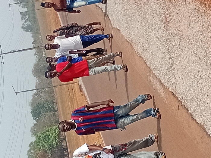 Eight men stand in a line along the edge of a road. Most are dressed in casual shirts and jeans. One wears a striped jersey, and another is shirtless. They appear to be posing or waiting together. Power lines and trees are visible in the background. The roadside is gravelly, contrasting with the paved road they are standing on. The scene is captured in a slightly skewed perspective.