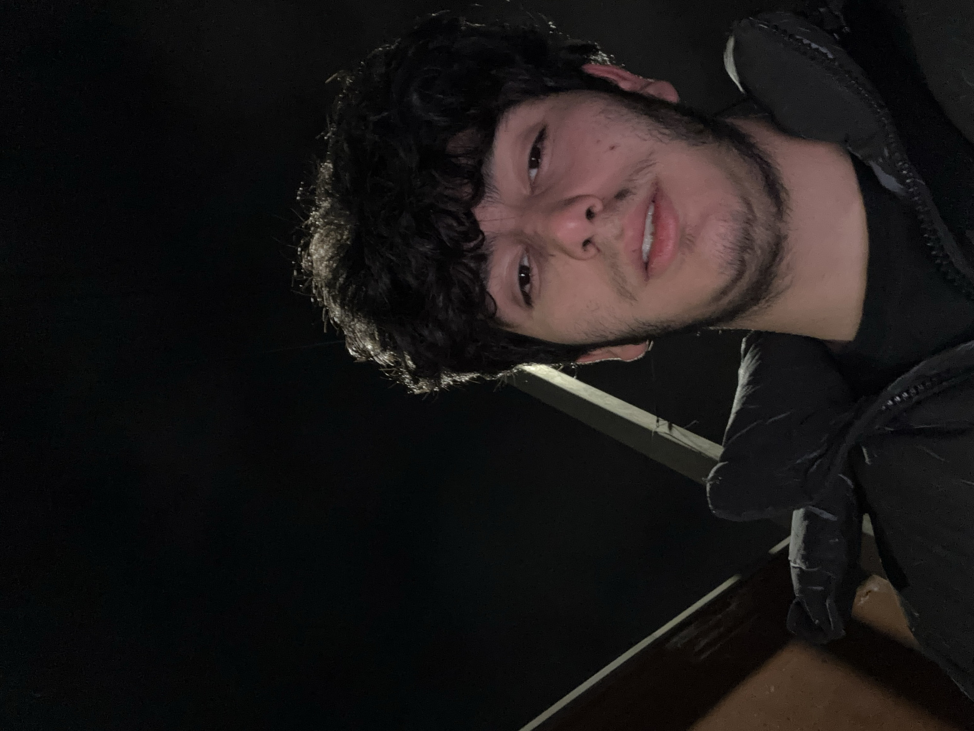 A low-angle photograph shows a young man with dark, curly hair and stubble. He is wearing a dark jacket. The background is mostly black, suggesting it is night. A metal structure is partially visible behind him. The location cannot be determined as Yerevan, Armenia, based on the information provided.