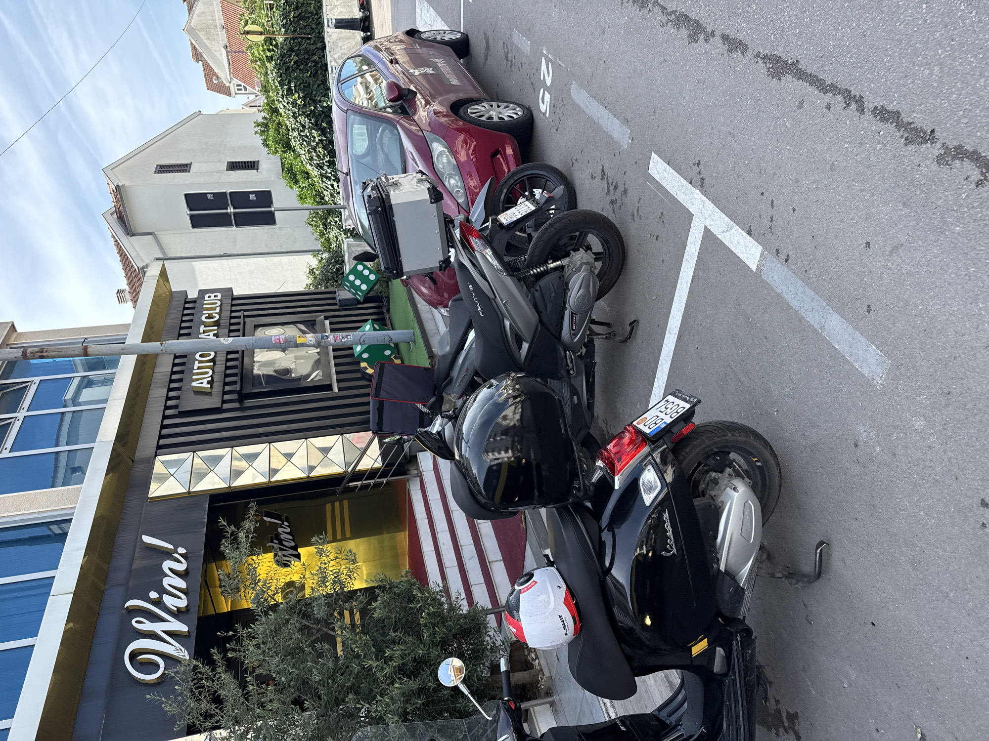 The image displays a sunny street scene in Budva, Montenegro, featuring a row of parked vehicles alongside commercial and residential buildings. The perspective is slightly elevated, looking down a street delineated by white parking lines.
In the foreground and mid-ground, several scooters are parked on the right side of the road. The closest is a black Vespa with a black helmet resting on its seat and a white, red, and black LS2 helmet hanging from the side. Its license plate, "BD 7938," clearly indicates Budva. Behind it, another black scooter with a silver top box is visible, with a license plate partially showing "BD 07-30." Further down, a red four-door hatchback car, possibly a Peugeot 207, is also parked. The number "25" is painted on the asphalt near the car.
On the left side of the street, a modern building features a prominent "Win!" sign in white script on a dark facade with gold metallic accents. Above it, another sign reads "AUTOMAT CLUB." Large green cubes, resembling dice, are placed near the building's entrance, consistent with a gaming establishment. Behind this modern structure, an older, light-colored residential building with a tiled roof and shuttered windows is visible under a clear, bright blue sky with a few wispy clouds. Green bushes and small trees line the sidewalk, adding to the urban landscape. The scene depicts a typical daytime street with no people visible.