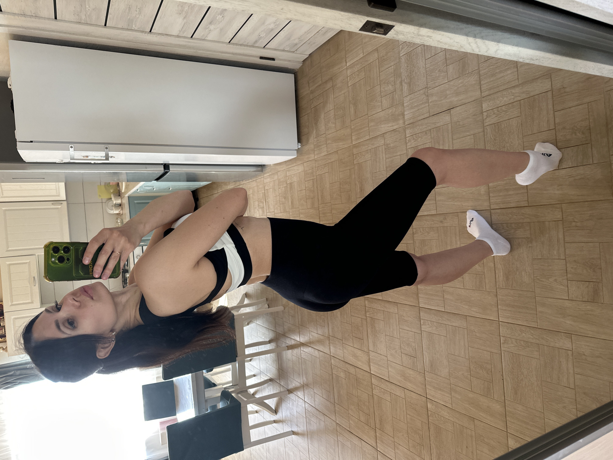 A person is taking a selfie in what appears to be a dining area or kitchen. The individual, a woman, is wearing a black and white sports bra and black leggings, with white socks on her feet. She is holding a smartphone, which has a green case, in her right hand, facing the mirror to capture the image. Her hair is long and dark, and she is looking at her reflection in the mirror.
The background includes a kitchen or dining room setting. There is a white refrigerator visible on the left side of the frame, along with kitchen cabinets and countertops. A dining table with chairs is also present behind her. The floor is covered with tiles in a light brown, wood-like pattern. The lighting appears to be natural, with sunlight streaming in from a window near the dining table.