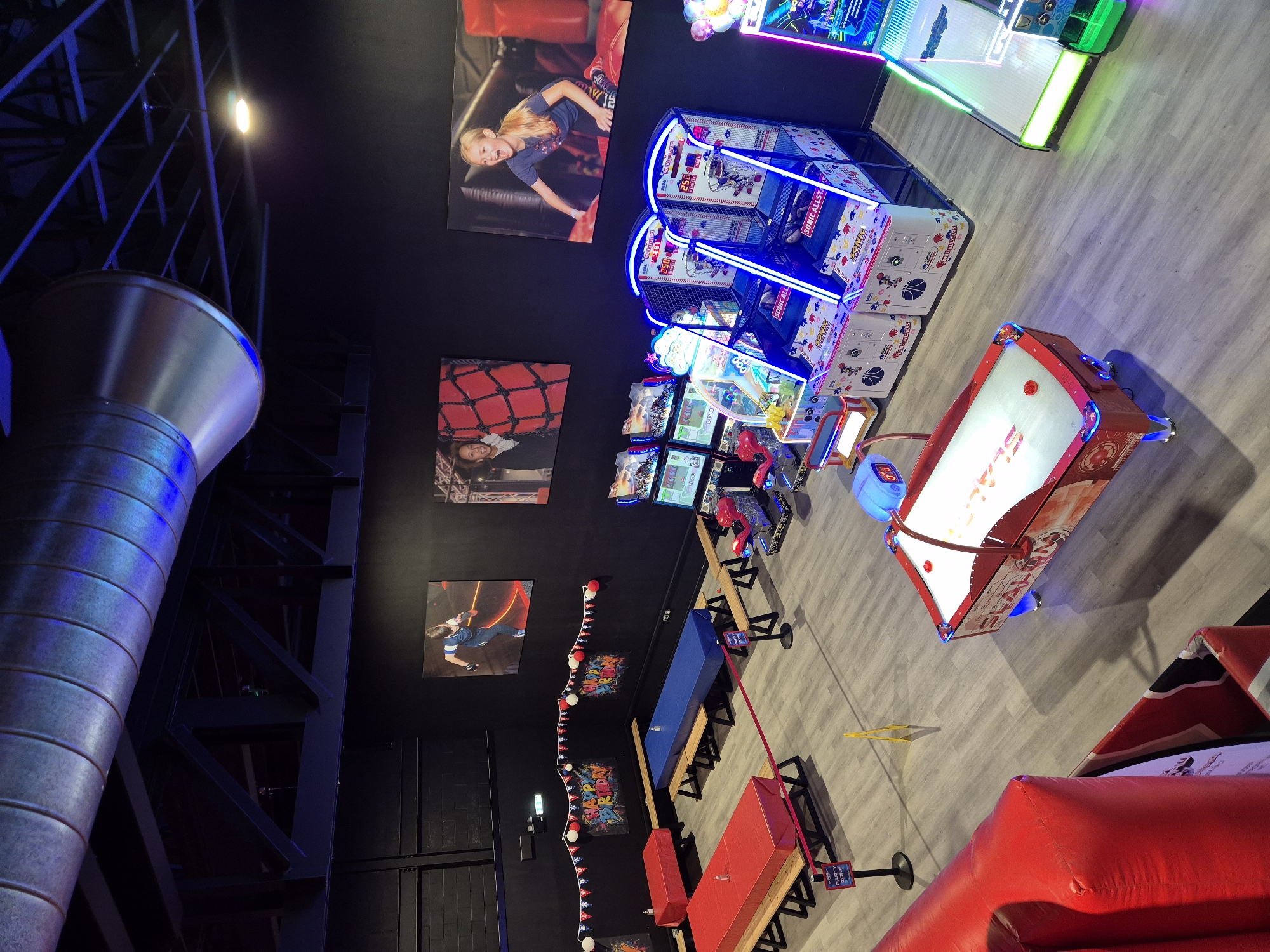 The image depicts the interior of a recreational facility, likely an arcade or entertainment center in Chelmsford, United Kingdom. The walls are dark, adorned with large pictures or advertisements related to the center. Multiple arcade games, some with neon lighting, are visible, including basketball hoops, and others with screens. There is an air hockey table. Tables and benches with red and blue covers are visible. There is a large metal structure in the foreground, likely part of the building's architecture. A yellow caution sign on the floor is present.