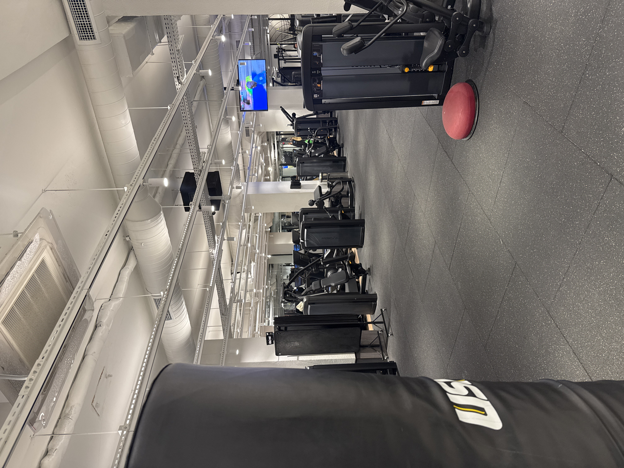 This is a photograph of the interior of a modern gym. The gym has a dark gray rubberized floor and numerous pieces of black gym equipment, including weight machines and a large punching bag on the left side of the frame. The ceiling is white and features exposed ductwork, lighting fixtures, and a large air conditioning unit. A flat-screen television is mounted high on the wall in the background, displaying a sports broadcast. The overall impression is one of a clean, well-lit, and functional fitness facility. The image does not provide any specific visual cues to determine its location within Kadıköy, Turkey.