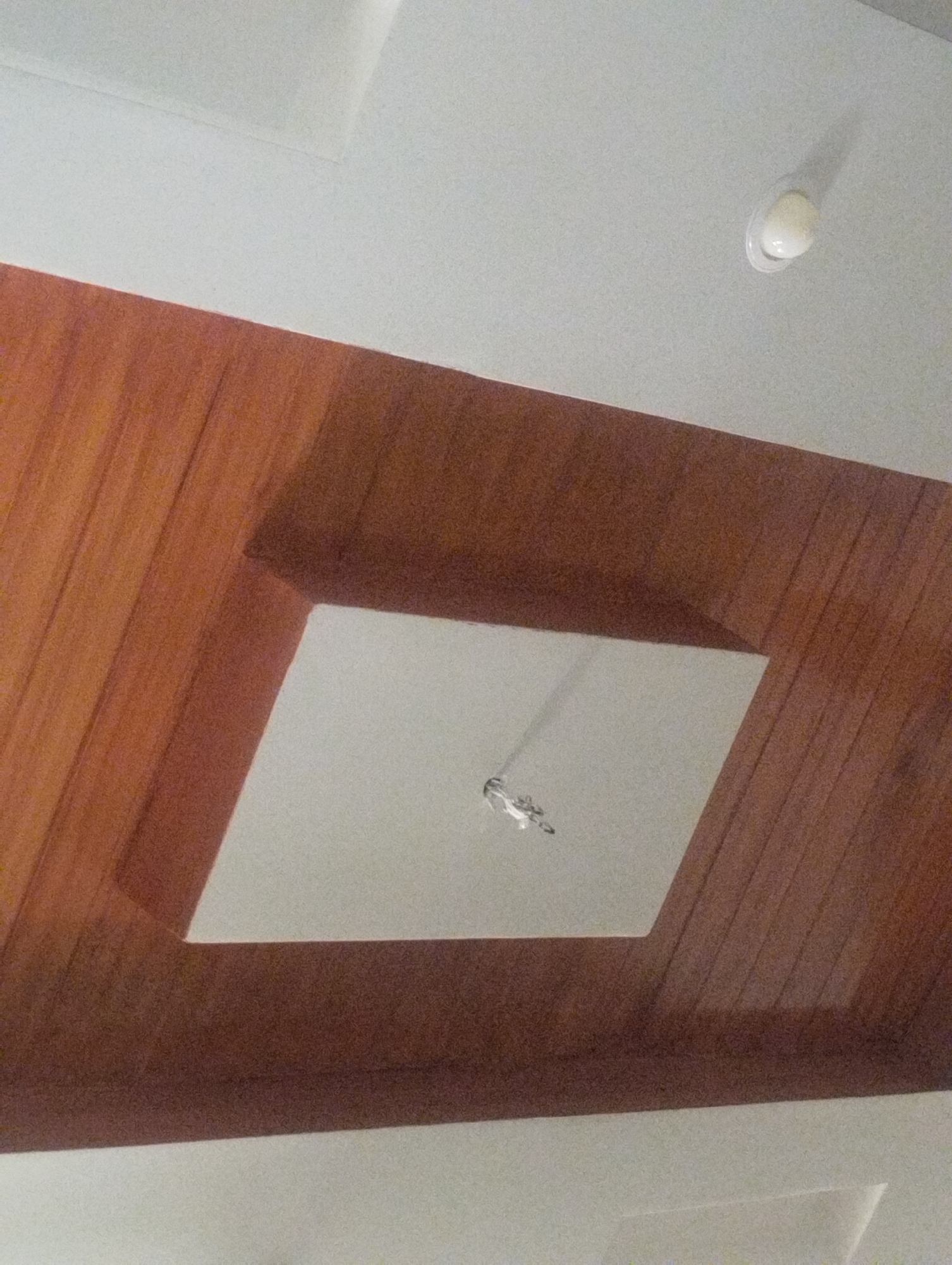 An indoor shot captures a portion of a ceiling. The ceiling features a recessed area with a light fixture. The main ceiling surface is white, and the recessed area is also white and square-shaped, with a small, clear light fixture positioned in its center. Surrounding the white recessed area is a wooden paneling that appears to be stained in a warm, reddish-brown hue. The wooden panels run parallel to each other, creating a textured surface that contrasts with the smooth white areas of the ceiling. The lighting in the image is ambient, casting soft shadows and highlighting the different textures of the materials. The scene is still and devoid of any people or animals.