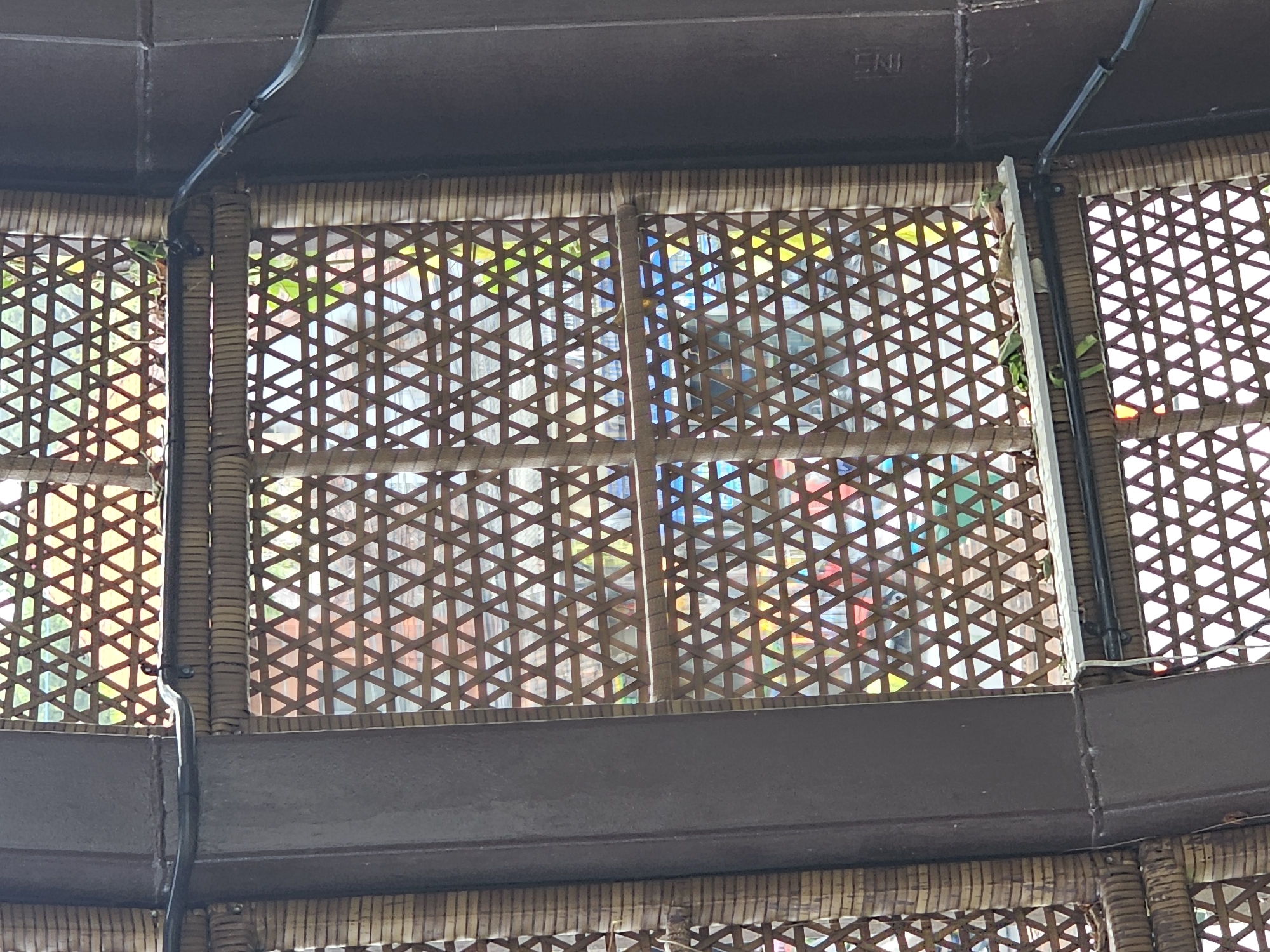 The image depicts an exterior view of a structure with a woven bamboo facade, likely a wall or partition, in Denpasar, Indonesia. The bamboo is arranged in a repeating triangular pattern, creating a latticework effect. Through the openings in the bamboo, colorful materials or lights are visible, suggesting an interior space with decorative elements. The structure appears to be supported by dark, vertical beams, and a black cable runs horizontally across the upper section of the woven facade. Some green foliage is also present, peeking through the bamboo openings in the middle section of the image. The overall impression is one of an architectural detail with an emphasis on natural materials and dappled light.