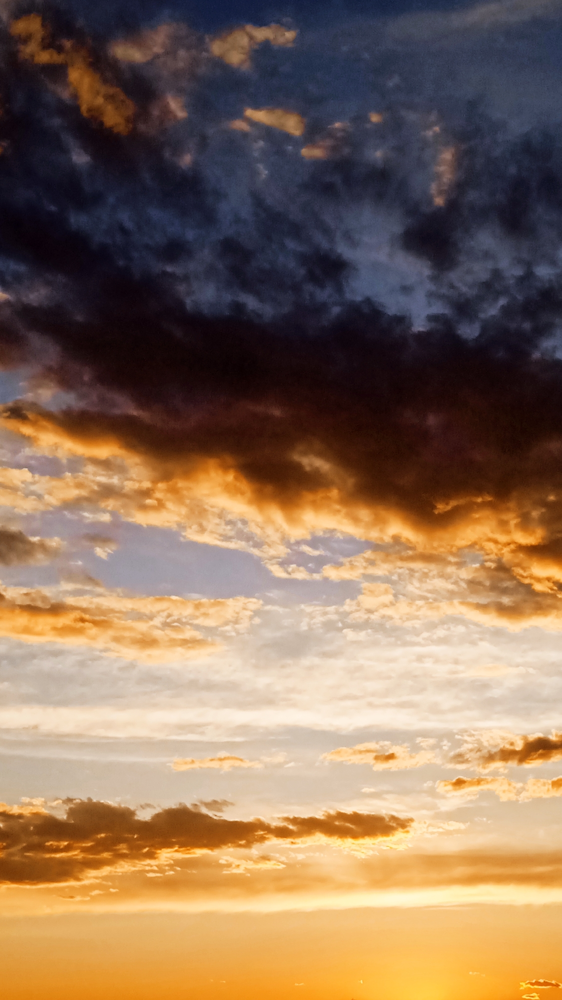The image depicts a dramatic sky filled with clouds during sunset or sunrise. The clouds range in color from deep blues and purples at the top to vibrant oranges and yellows towards the bottom, indicating the presence of the sun just below the horizon. The light from the sun illuminates the undersides of the clouds, creating a striking contrast between the shadowed and lit areas.
The scene is entirely focused on the sky, with no discernible objects or people. The cloud formations are varied, with some appearing wispy and scattered, while others are thicker and more defined, catching the light in bold strokes. The overall impression is one of awe and grandeur, characteristic of a vivid sunset or sunrise. The colors suggest a warm and potentially serene atmosphere, although the darker clouds at the top add a touch of drama. There is no visible text in the image.
