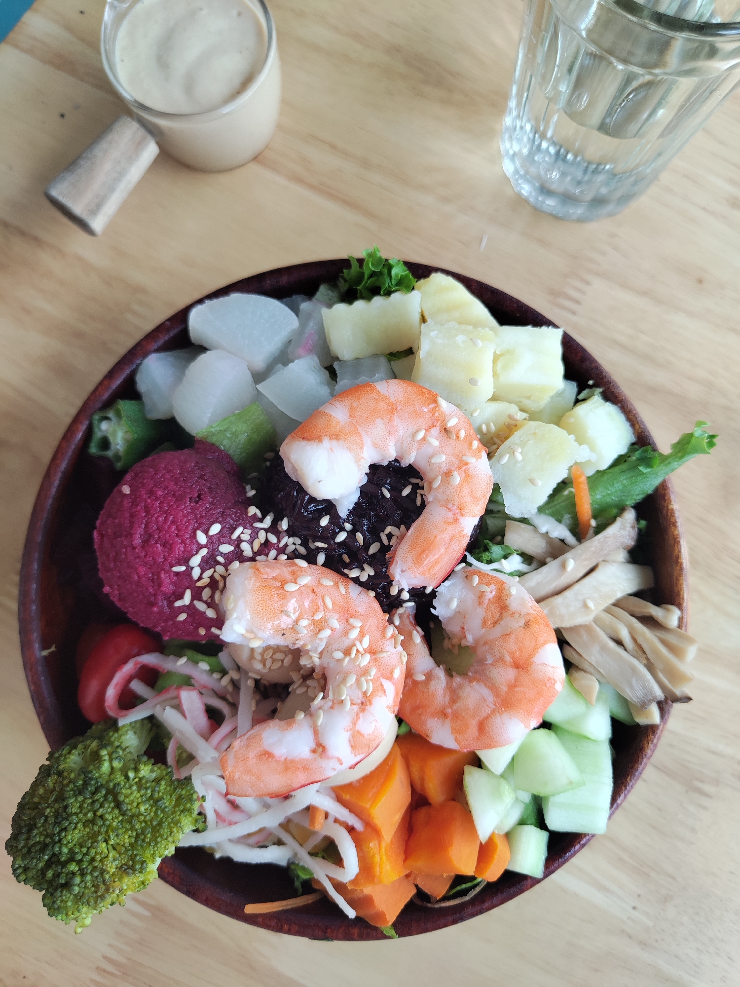 The image shows a wooden bowl containing a salad with various ingredients. These include three cooked shrimp sprinkled with sesame seeds, cubes of white radish, pineapple, chunks of carrot, cucumber, shredded imitation crab meat, a dark purple scoop (likely beetroot hummus), black rice, sliced mushrooms, broccoli, green vegetables, and a few small red tomatoes. To the upper left of the bowl is a small glass jug containing a light-colored dressing. To the upper right is a glass of clear liquid, presumably water. The bowl, jug, and glass are on a light-colored wooden table. The location is Da Lat, Vietnam.