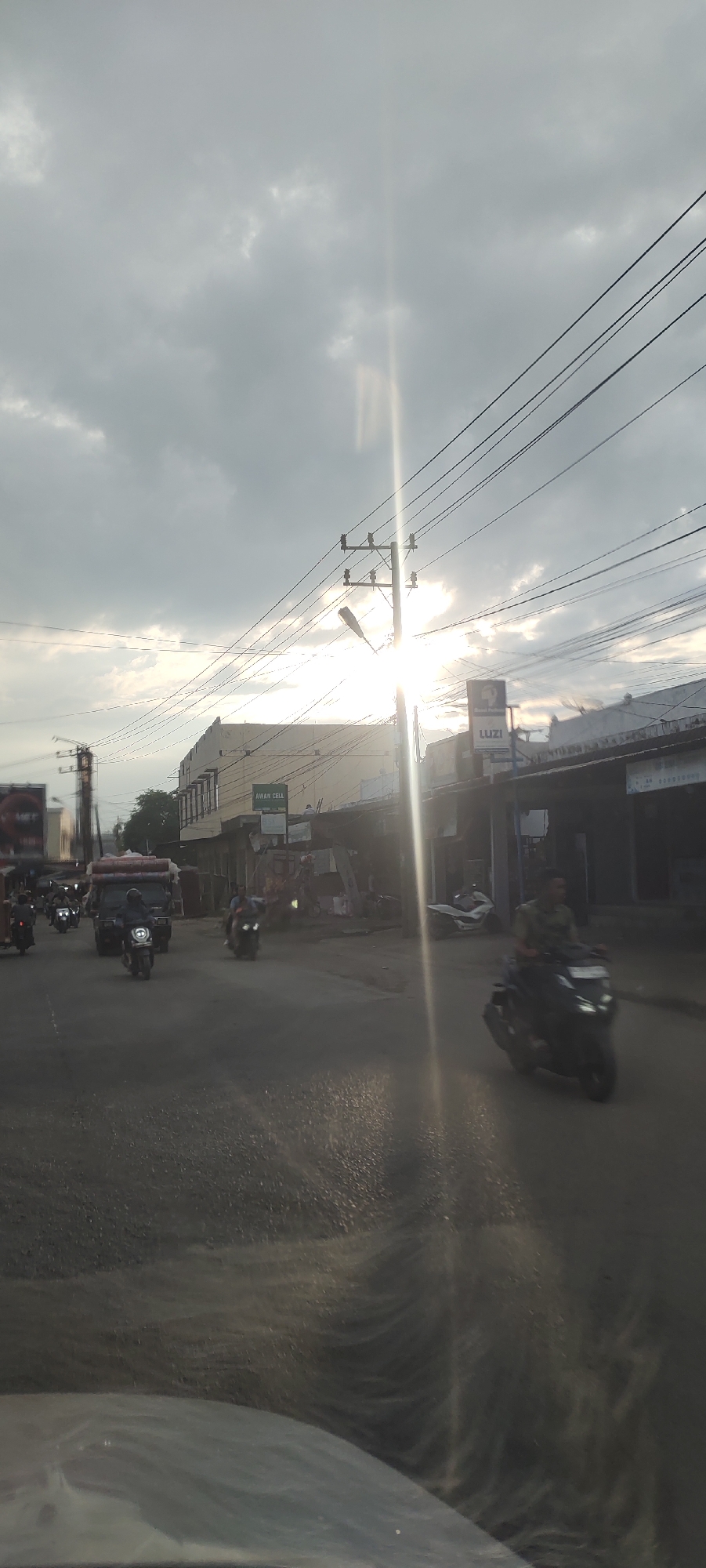 The image depicts an urban street scene in Keudegeudong, Indonesia, during what appears to be late afternoon or early evening, characterized by strong backlighting. The sky is mostly covered with scattered grey clouds, and intense sunlight projects a prominent lens flare and visible rays from the upper right side of the frame.
Multiple black power lines crisscross the upper and middle portions of the image, connecting to several utility poles. A street light fixture is mounted on the primary pole positioned towards the right of the center. On the left side of the street, a multi-story building, light in color, features a sign with "AWAN CELL" prominently displayed. Further down the street on the right, another commercial building has a sign that reads "LUZI."
The asphalt road surface in the foreground and midground exhibits a reflective quality, suggesting dampness or the effect of the low sun angle. Vehicular traffic includes numerous motorcycles and one small truck. A truck loaded with bulky items, possibly mattresses or textiles, is visible in the middle-left, preceding a person on a motorcycle. On the right side of the road, a person wearing a camouflage-patterned shirt is riding a dark-colored scooter, moving away from the viewer. Additional motorcycles and individuals are observed farther down the street. The perspective indicates the photograph was taken from inside a vehicle, with reflections visible along the bottom edge of the image.