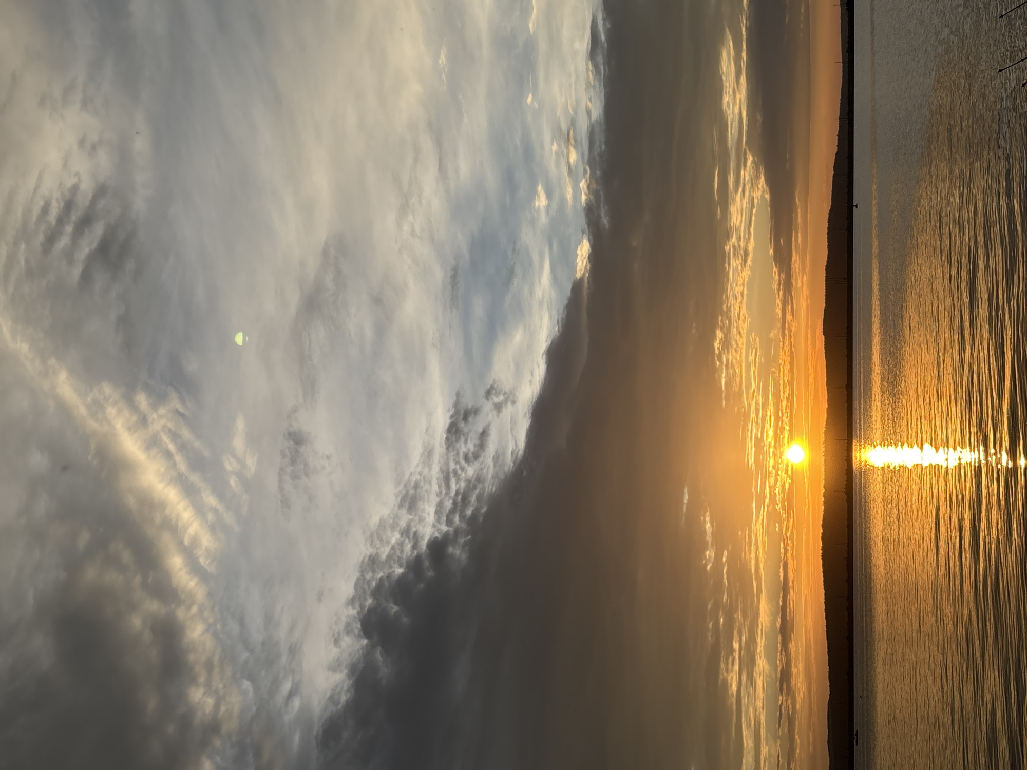 The image depicts a sunset over a body of water, likely a river or lake, as seen from the Chaikovsky Reservoir in Chaikovsky, Russia. The sun, a bright orange orb, is low on the horizon, casting a brilliant reflection on the rippling water. The sky is filled with dramatic clouds, ranging from dark gray to light, wispy formations, some illuminated by the setting sun. The foreground is dominated by the water, with a dark, silhouetted treeline or hilly landscape visible in the distance. Two small boats are faintly visible on the water. The overall scene is one of natural beauty and tranquility.