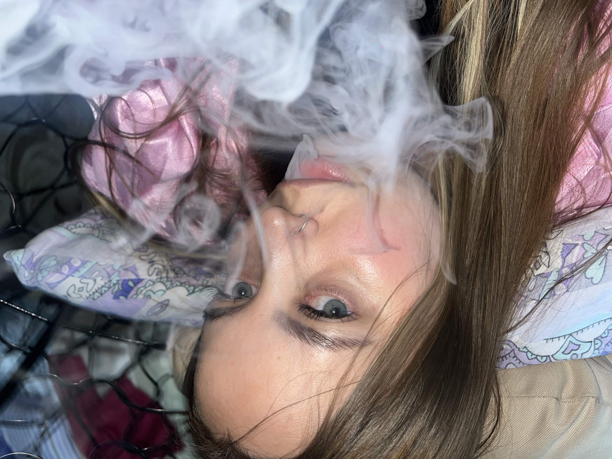 A young woman with brown hair and blue eyes is lying down, looking up at the camera. She has a nose ring in her left nostril. Smoke obscures part of her face. She is wearing a pink top and is lying on patterned pillows on a black wire rack. There is no evidence to support a location in Novopokrovskaya, Russia.