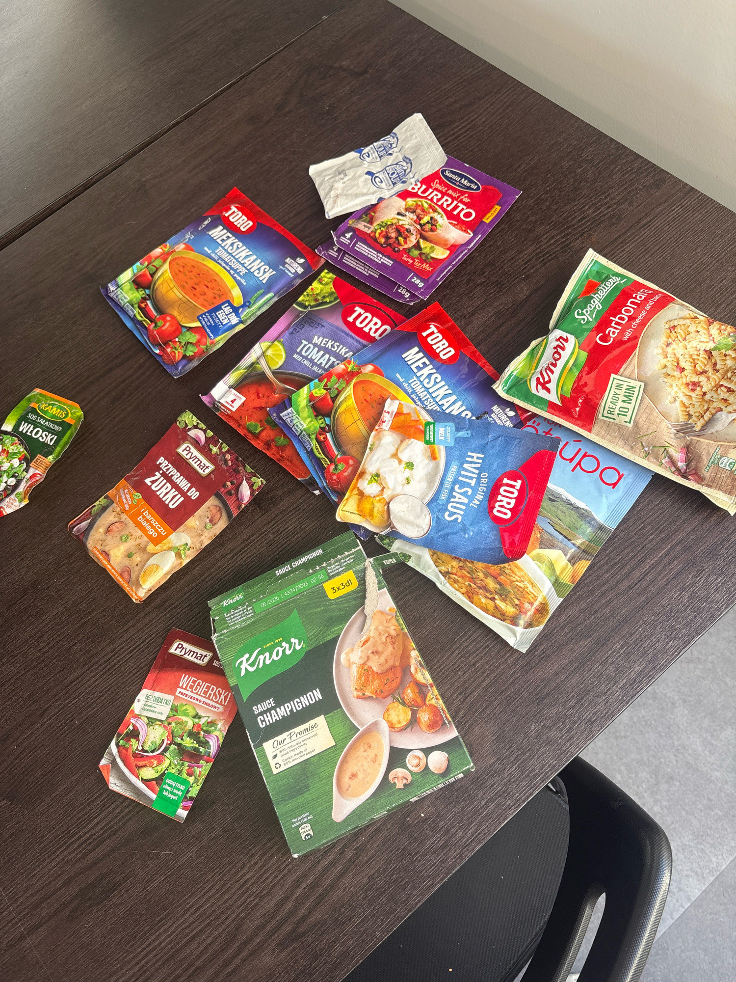 The image shows a dark brown wooden table with various food seasoning packets and boxes scattered across it. The items include TORO brand soup mixes, SANTA MARIA brand burrito spice mix, SPAGHETTERIA brand carbonara, Knorr brand mushroom sauce, and other Polish brand seasoning packets. The lighting suggests an indoor setting, possibly a home kitchen or a store shelf. The scene does not contain any elements that would indicate its location in Reykjavik, Iceland. There are no people or specific actions depicted.