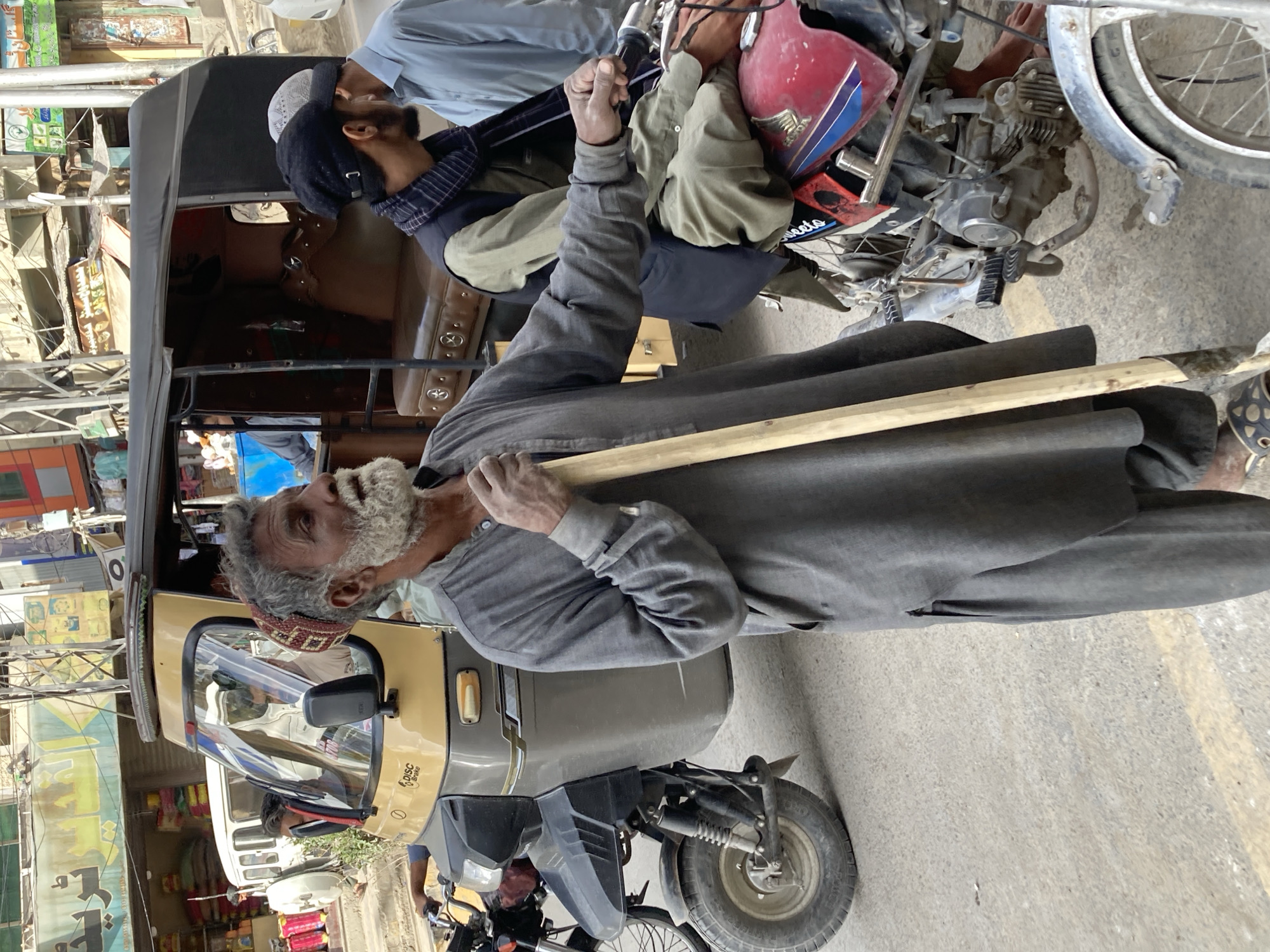 An elderly man with a white beard wearing a grey tunic and a red and brown cap stands in front of a yellow and black auto rickshaw (tuk-tuk) in what appears to be a street. He is holding a long, light-colored wooden pole. Another man wearing a light blue shirt and a black cap with white embroidery is nearby, leaning over a motorcycle with a red and blue tank, possibly attending to it. The backdrop features buildings and shops, typical of a city street. The scene is likely located in Quetta City Tehsil, Pakistan.