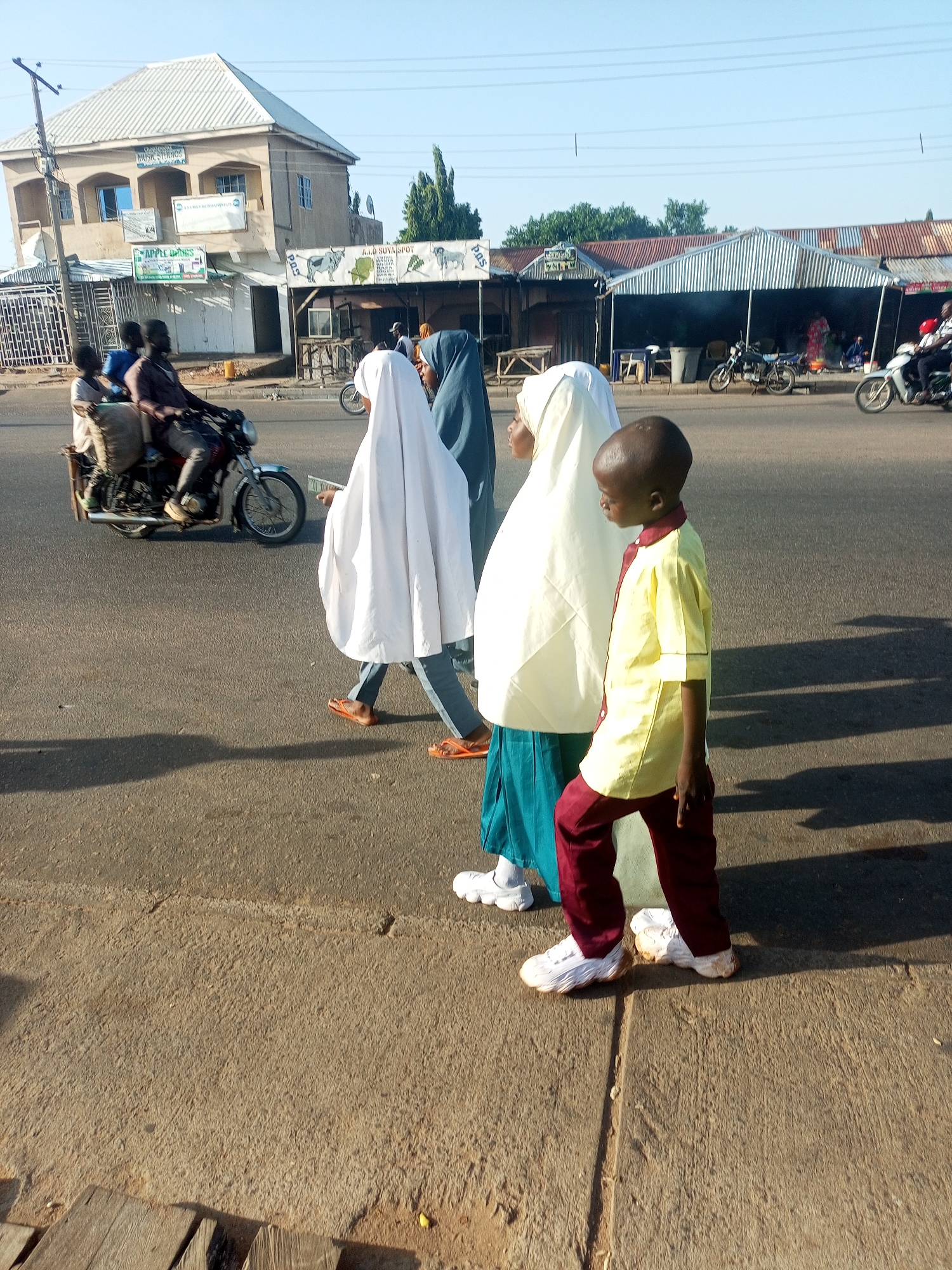In Tumfure, Nigeria, this image captures a street scene with several individuals and elements of urban life. In the foreground, a young boy wearing a yellow shirt and maroon pants walks on a paved surface, casting a shadow. Behind him, three individuals, likely women, are walking. Two are covered in white hijabs, and one is in a blue hijab. One of the women in white is wearing orange flip-flops. To their left, a motorcycle with a rider and two passengers is moving on the asphalt road. The passengers appear to be carrying a large, bagged item. In the background, buildings line the street. One building, a multi-story structure with a white roof, has signs indicating "MUSIC STUDIOS" and "EXTRA MINING INVESTMENT LTD." Adjacent to it are market stalls with corrugated metal roofs. Further down the road, other motorcycles and individuals are visible. The sky is clear and bright, indicating daylight. The overall scene depicts a typical day in this Nigerian city, with people engaged in daily activities on the street.