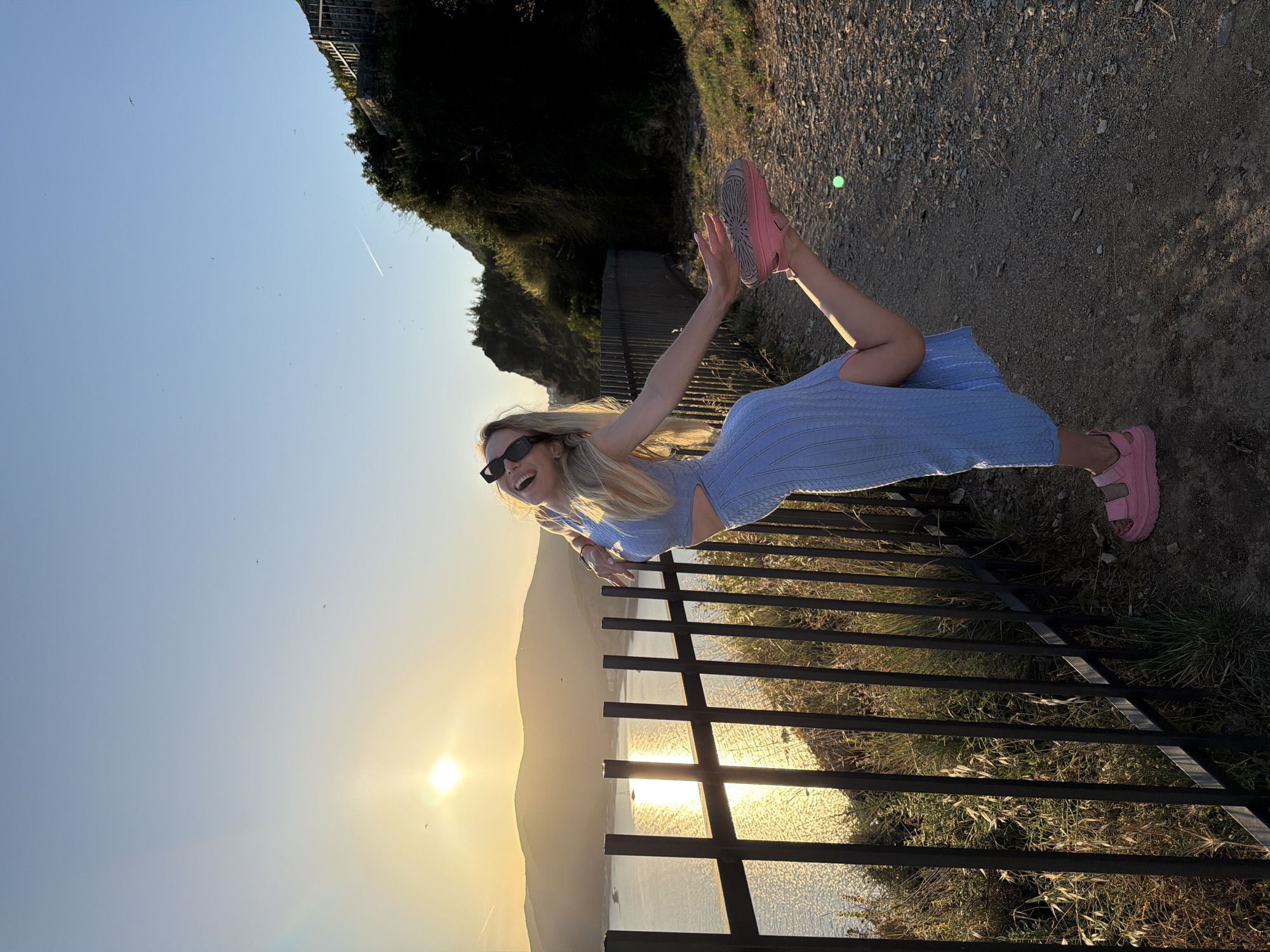 A blonde woman, wearing a light blue knit two-piece ensemble consisting of a crop top and a midi skirt with a side slit, black sunglasses, and chunky pink platform sandals, is captured in a playful pose on a dirt path. She is balancing on her left leg, with her right leg bent and raised, holding the removed pink sandal from her right foot in her hand. Her head is tilted back, mouth open in an expression of laughter, and her arms are extended outwards. The woman stands next to a black metal fence running along the edge of a cliff or elevated path.
The scene is illuminated by the golden light of a setting sun, positioned low on the horizon over a large body of water, likely the Adriatic Sea, which reflects the sun's brilliance. Distant mountains or hills rise from the opposite side of the water. The sky above is clear, transitioning from a light blue to a warm yellow gradient near the sun, with several small birds and a faint airplane contrail visible. To the woman's right, the path continues alongside a slope covered in sparse vegetation and some larger bushes. This coastal viewpoint is located in the city of Budva, Montenegro, characteristic of its scenic Riviera overlooking the sea and mountains.