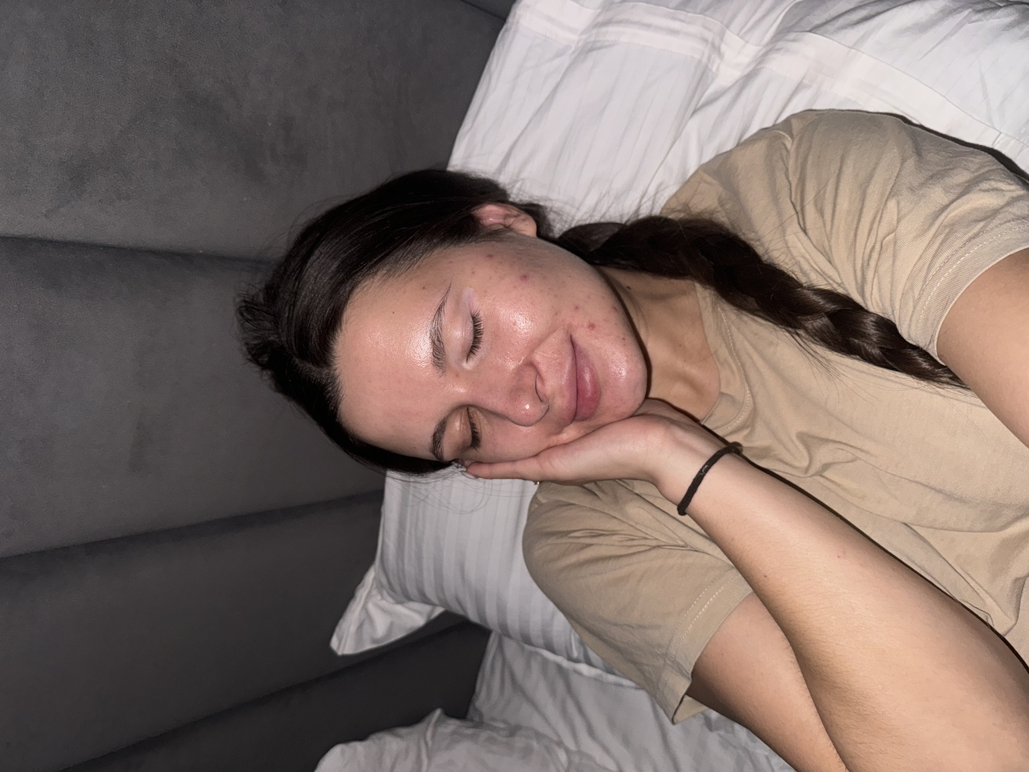 A young woman is asleep on a bed. She is lying on her side with her head resting on her hand. She has dark hair that is braided. She is wearing a tan T-shirt and a black bracelet. The bed has white sheets and pillows. The background is a gray upholstered headboard. The location is Akulovo (Gp Kubinka), Russia.