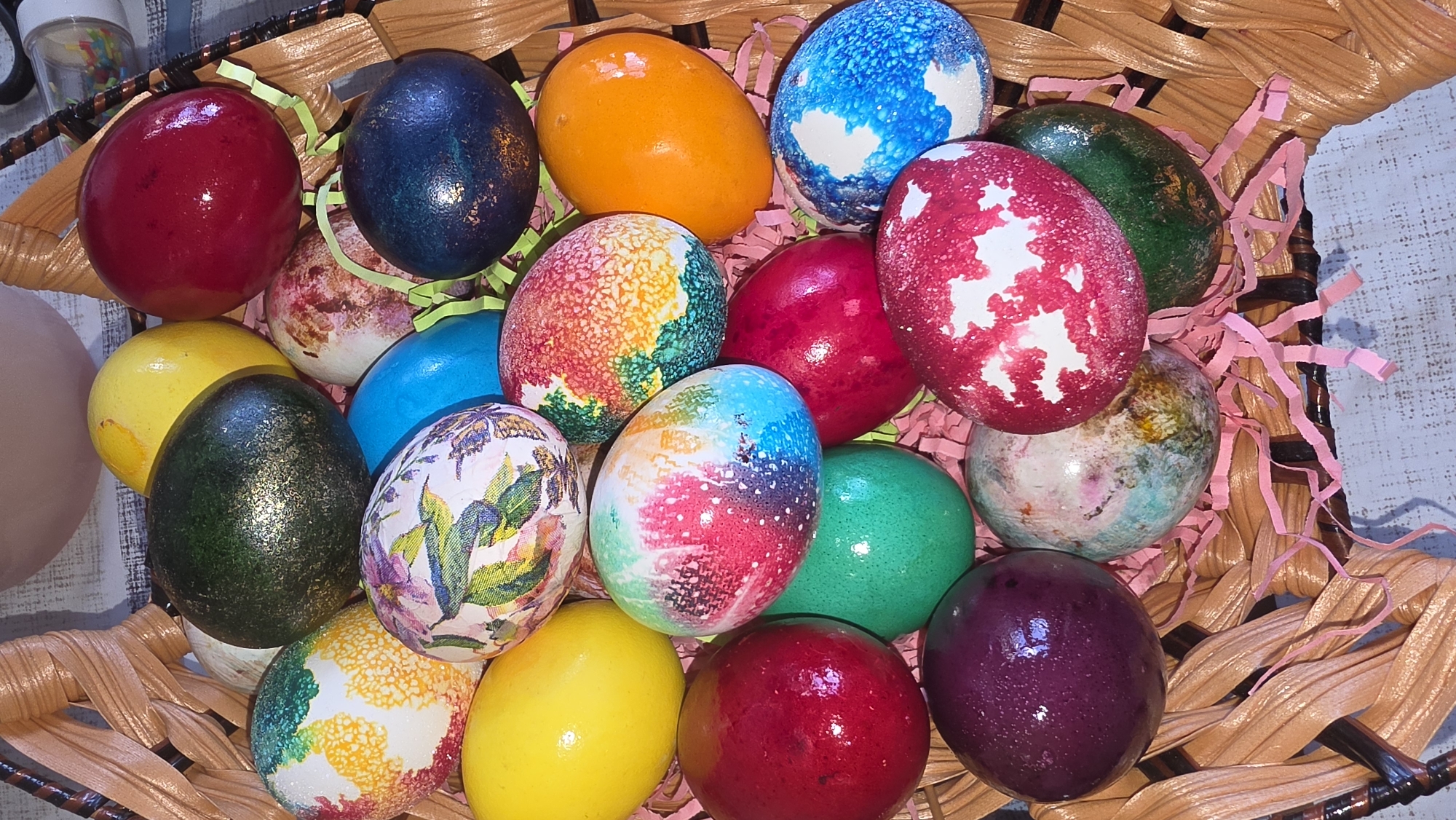 A close-up, eye-level shot depicts a collection of brightly colored and uniquely decorated Easter eggs nestled in a woven basket lined with pink paper shred filler. The eggs vary in color and design, including solid colors like red, yellow, orange, blue, green, and purple. Some eggs feature intricate patterns, such as multicolored speckles, floral designs, and speckled marble effects. The basket is a light brown, woven material. The location is not evident from the image.