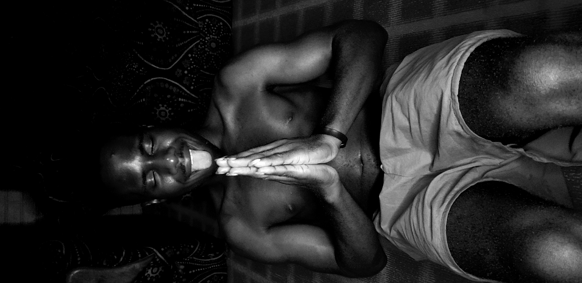 The image shows a dark-skinned man lying on his back, possibly on a bed, in what appears to be an indoor setting, presumably in Narayi, Nigeria. He is wearing shorts and has his hands pressed together, extended over his body. His tongue is sticking out. The environment is dimly lit, with a patterned surface visible in the background.