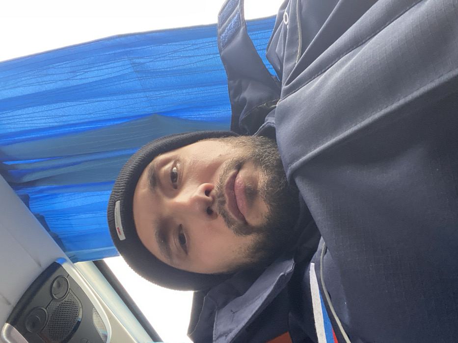 A close-up, slightly upward-angled image depicts a male individual inside a passenger vehicle in Luzhitsy, Russia. The man, with a short beard and mustache, is wearing a black beanie with a visible white "3M Thinsulate" label on the front and a dark blue jacket or uniform with a red, white, and blue stripe on the upper left shoulder area. He maintains a neutral expression and looks directly at the camera. The background features a blue pleated curtain covering a window on the right side, revealing a bright, overcast sky beyond. To the upper left, an overhead compartment with integrated lights and vents is visible, consistent with the interior of a bus or train.