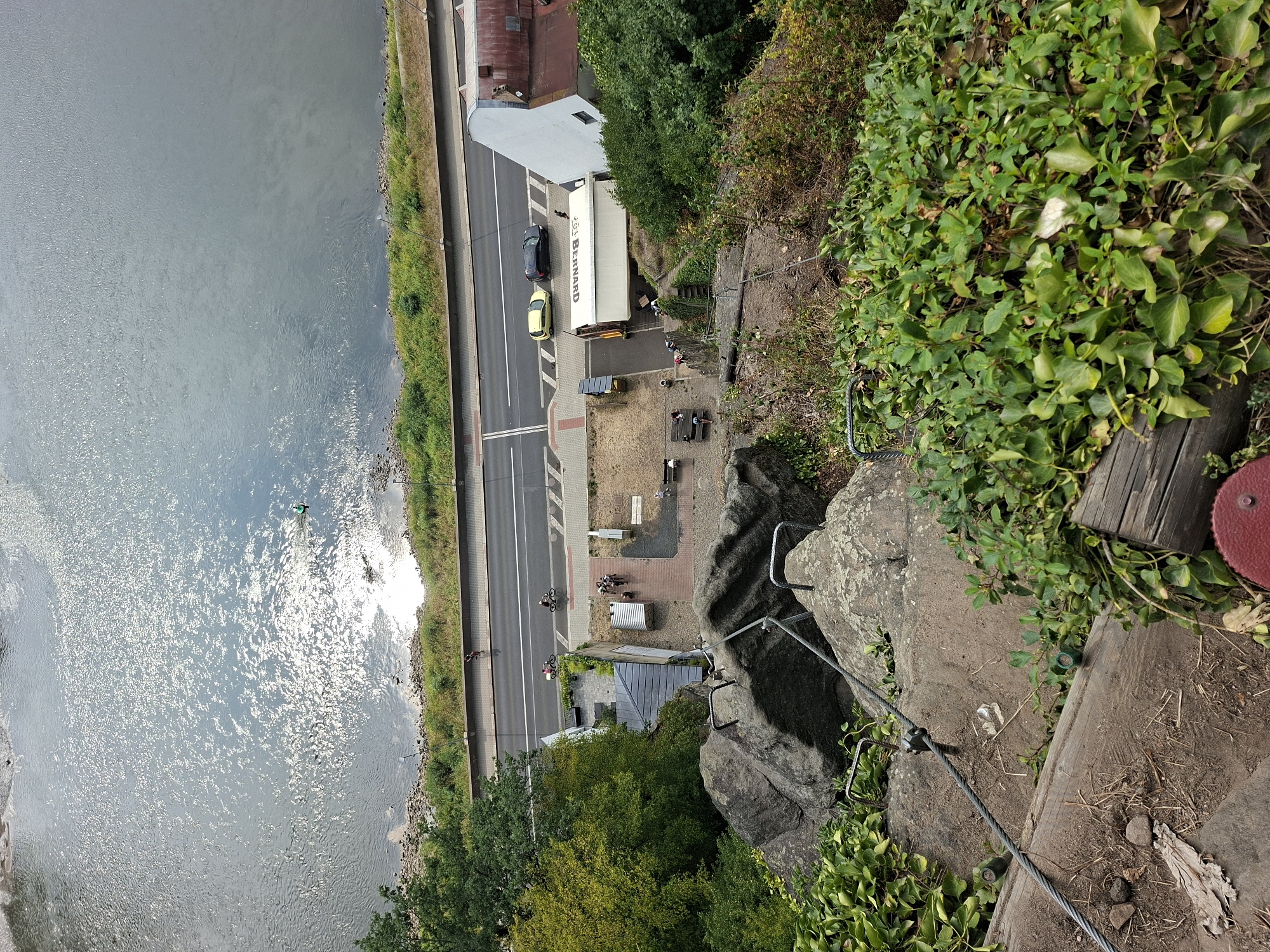 This image captures a high-angle view of a steep, rocky incline overlooking the Elbe River in Děčín, Czechia. The foreground shows a weathered wooden structure and dense green foliage. A steel cable and metal handrails are bolted into the rock face, indicating a hiking or climbing path descending towards a paved area below.
On the paved area, a group of people are gathered near a white truck with "BERNARD" printed on its side. Two yellow and black cars are parked on an adjacent road. Further beyond the road, the Elbe River is visible, reflecting the sunlight. A small, green-ringed object, possibly a buoys or a person in a life jacket, is in the river. A lone cyclist is seen on the road in the distance. The environment appears to be a scenic overlook or a recreational area.