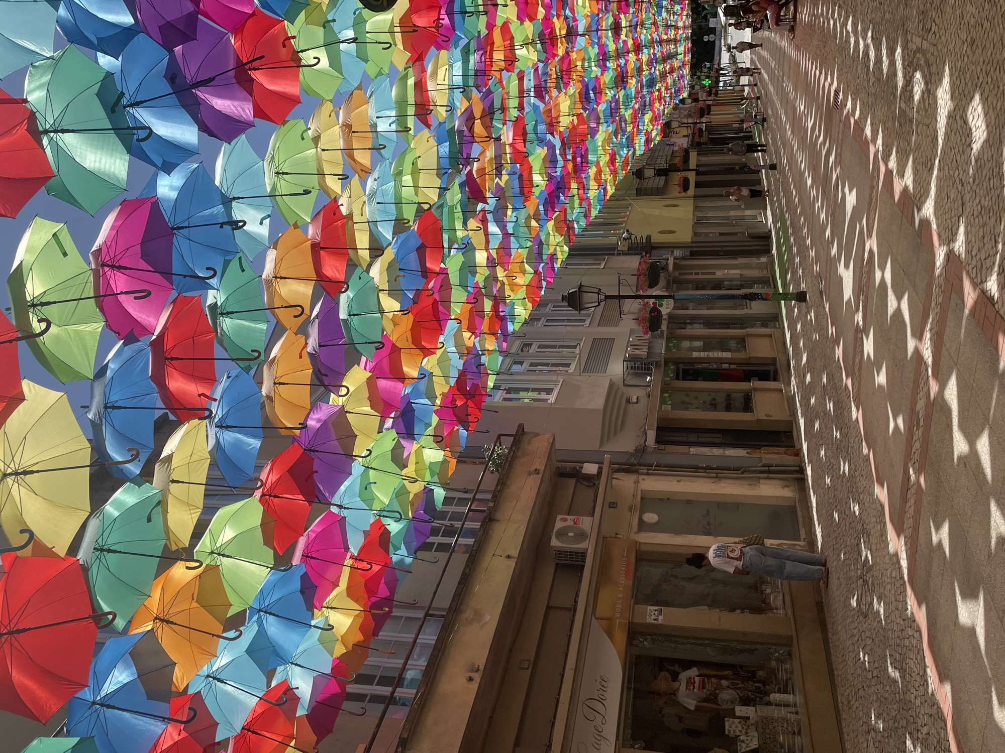 This media file captures an overhead view of a street lined with shops and buildings, adorned with a vibrant canopy of colorful open umbrellas. The umbrellas are suspended at varying heights, creating a striking visual spectacle that stretches down the street. The scene is bathed in bright sunlight, casting intricate star-shaped shadows from the umbrellas onto the cobblestone pavement below.
Several people are visible, walking along the street and interacting with the environment. One person, wearing a white t-shirt and blue jeans, is walking away from the camera on the right side of the frame, with their back to the viewer. Other individuals are seen in the distance, further down the street, some appearing to be patrons of the shops.
The shops lining the street feature large glass windows, displaying various goods and offering glimpses into their interiors. Some windows have signage, including a visible sign that reads "MODA & ACESSÓRIOS" and another that appears to say "Capa Dorée". The architecture of the buildings suggests an urban European setting.
The overall impression is one of a lively, sun-drenched street, transformed by the artistic installation of colorful umbrellas. The bright lighting and the abundance of color evoke a cheerful and festive atmosphere. The presence of people indicates that this is a public space, likely a shopping area or a pedestrian zone, experiencing a typical day. The time of day is clearly daytime, with the strong shadows suggesting the sun is high in the sky. The location is Águeda, Portugal, known for its "Umbrella Sky Project."